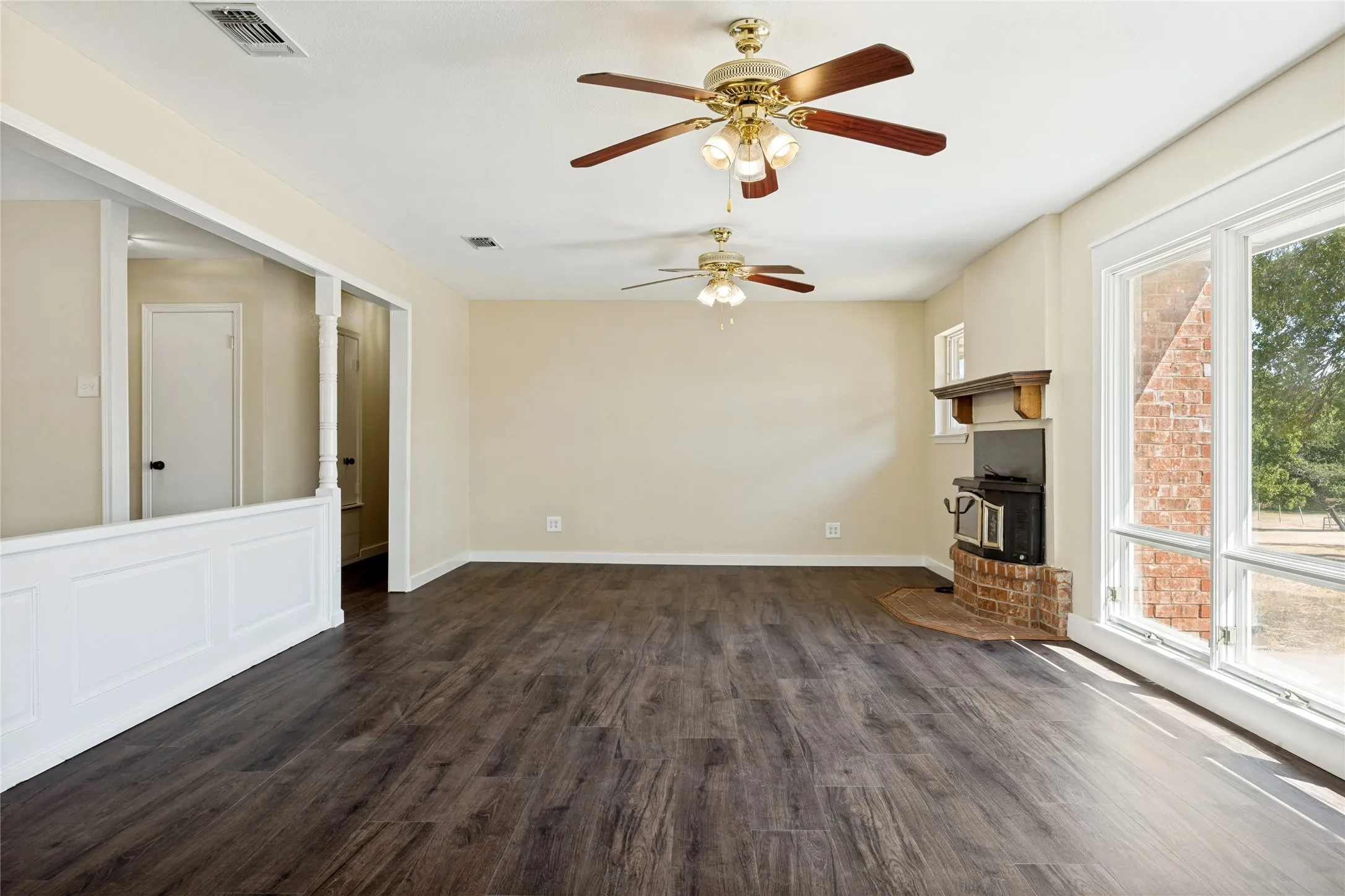 Wood-finish flooring, multiple ceiling fans with integrated lighting, brick fireplace surround with a wood mantel, and large picture windows