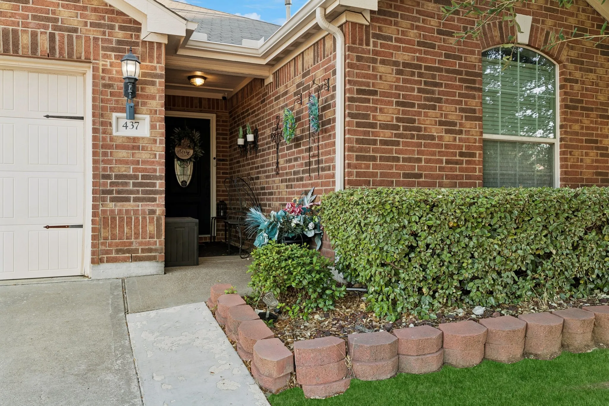 Brick facade with an arched window, white garage door, concrete pathway, and landscaping with a hedge