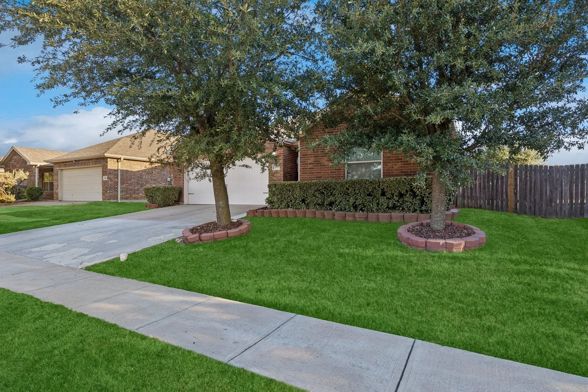 Brick exterior, attached two-car garage, concrete driveway, sidewalk access, and mature trees with landscaped beds