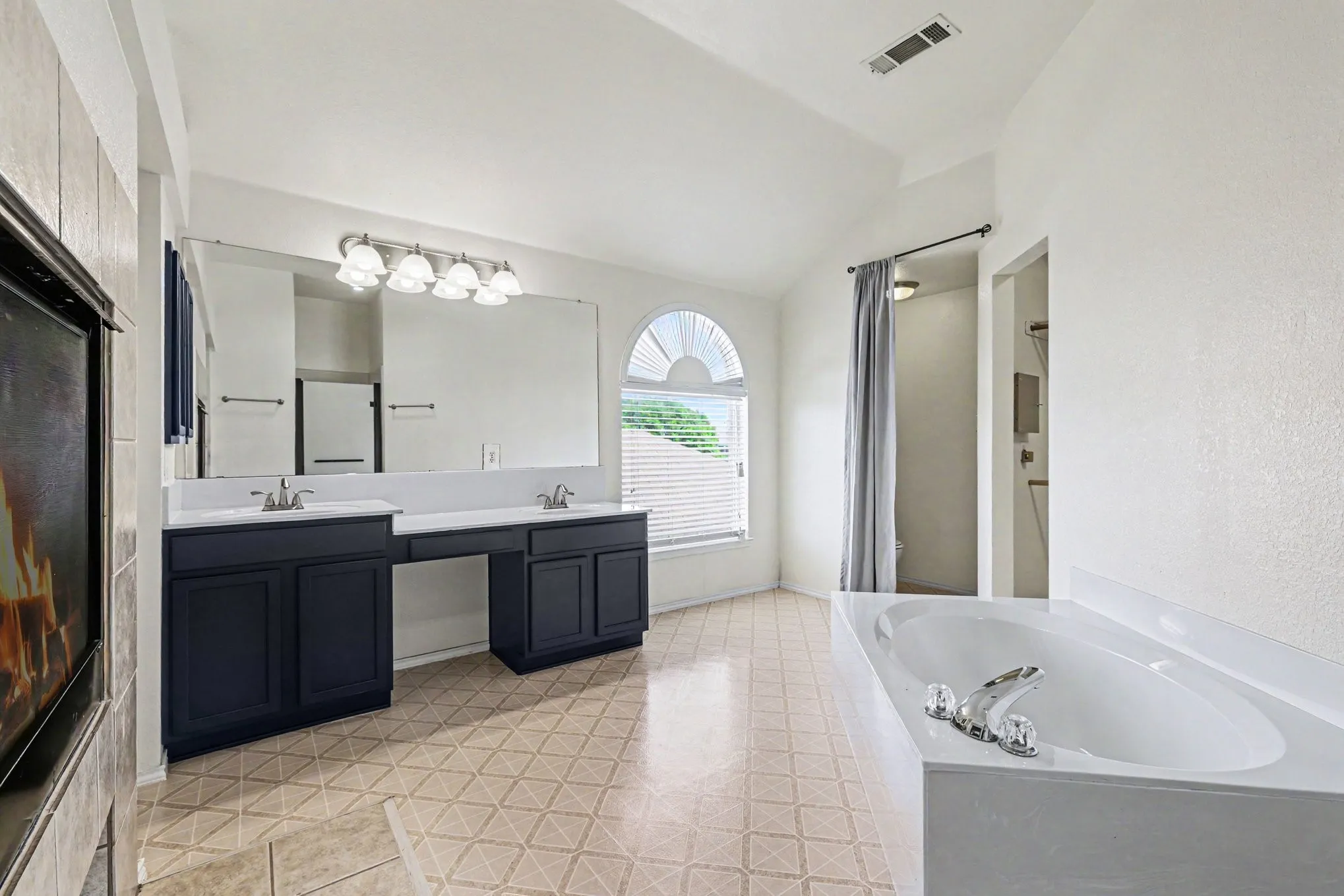 Vaulted ceiling bathroom featuring dual vanities, an arched window with blinds, a built-in soaking tub, and a separate shower enclosure