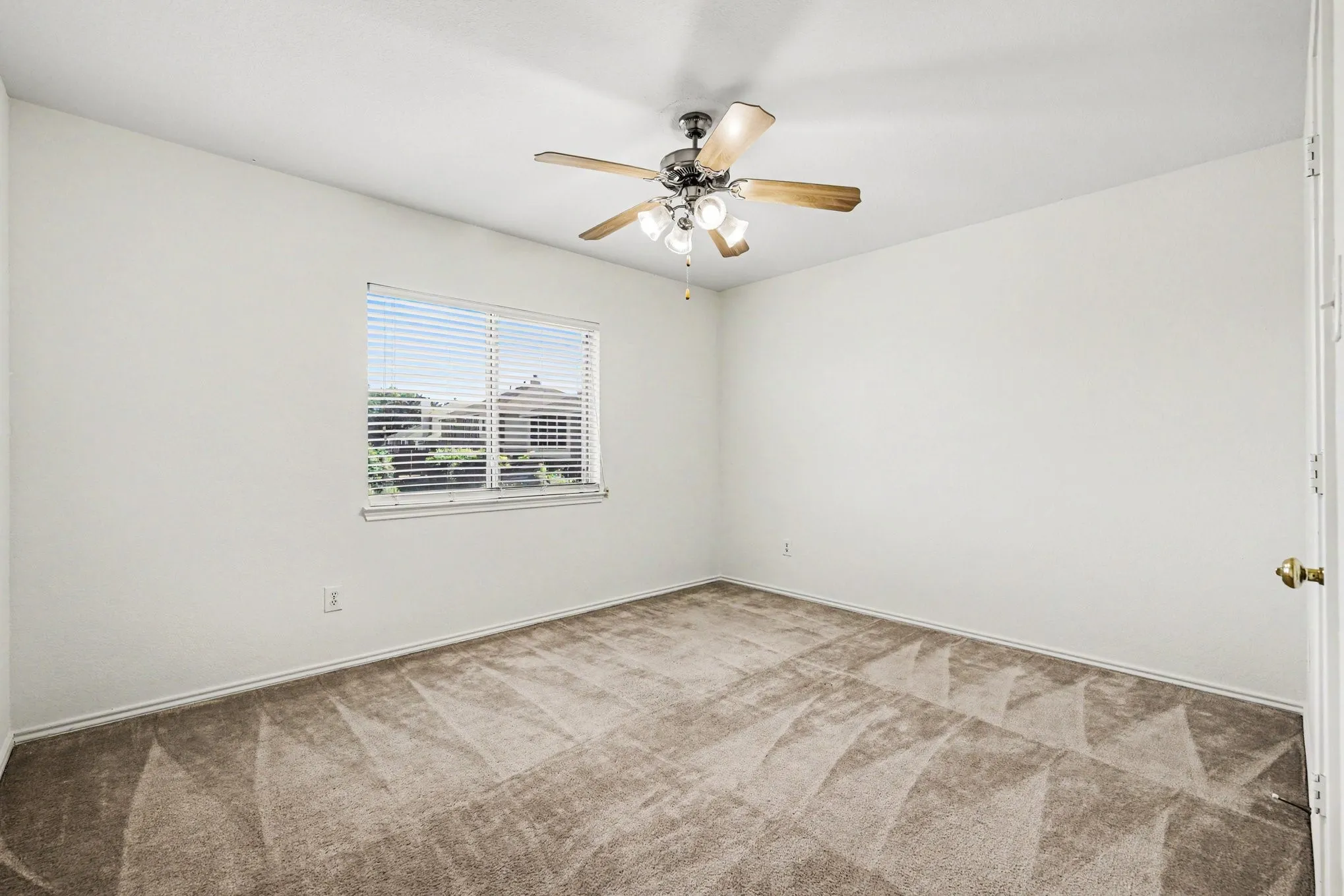 Neutral tone wall paint, textured carpet flooring, window with blinds, ceiling fan with integrated lighting, and a brass-toned door handle