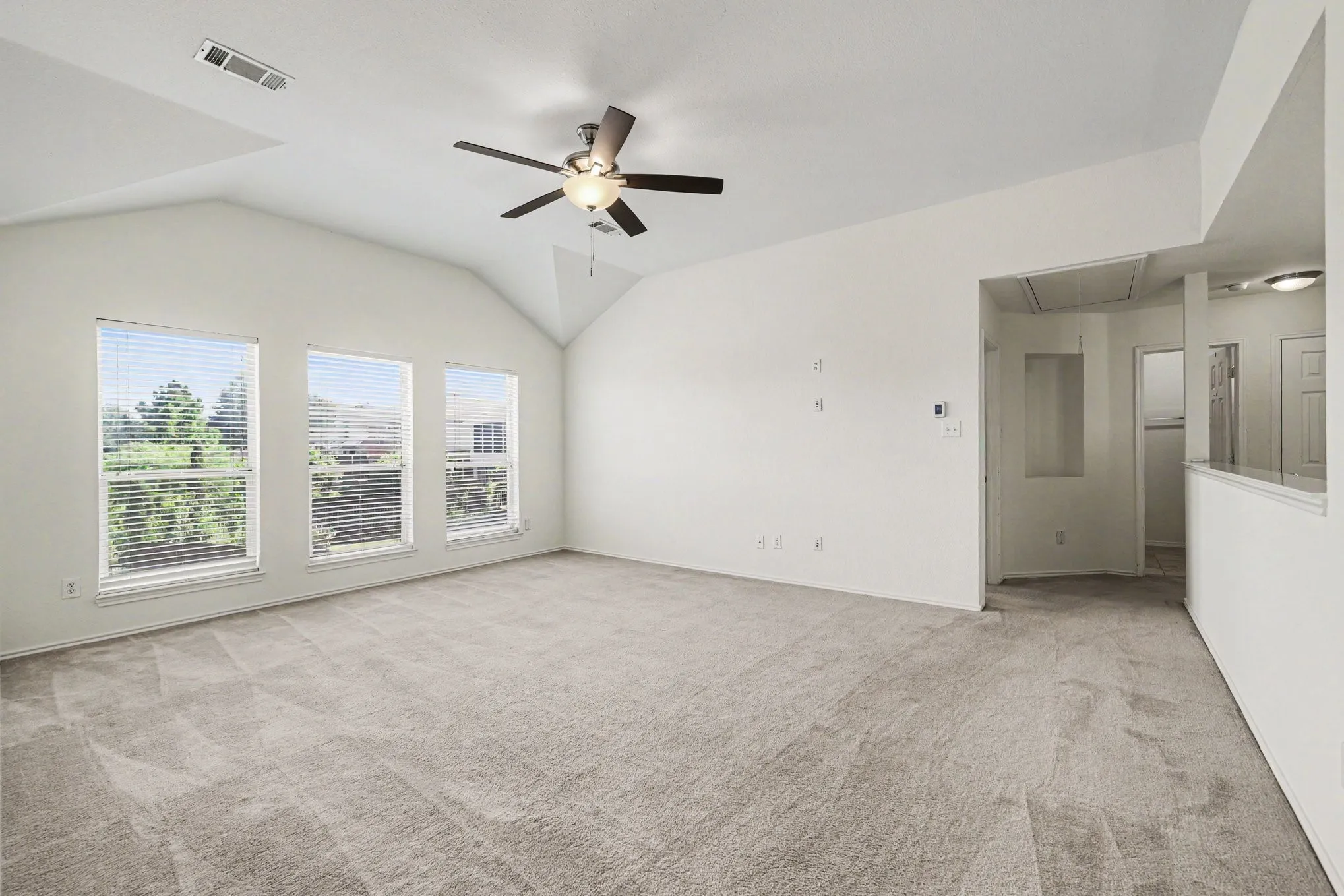 Vaulted ceiling with a ceiling fan, three windows with blinds, light-toned carpet, and an open half-wall partition