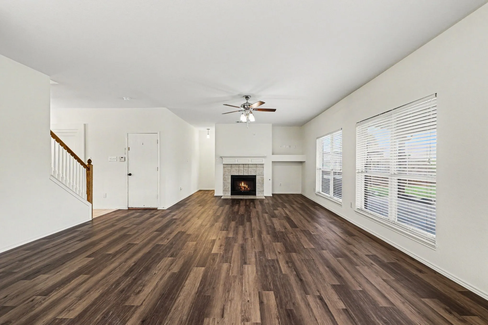 Spacious interior featuring wood-finish flooring, a fireplace with a mantel, multiple windows with blinds, a ceiling fan, and a staircase with a wooden handrail