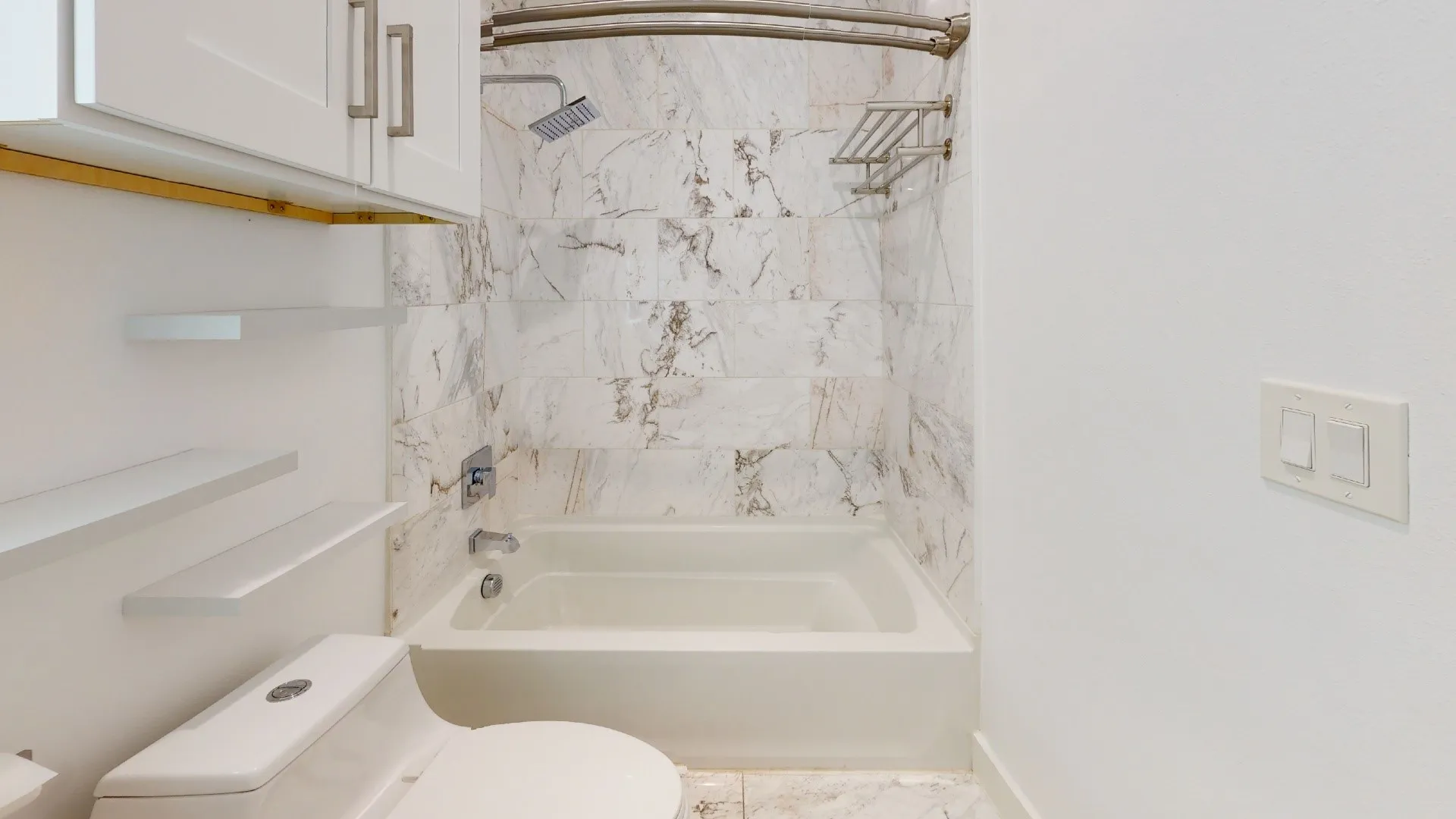 Bathtub with a shower, marble-style wall tiling, and white floating shelves