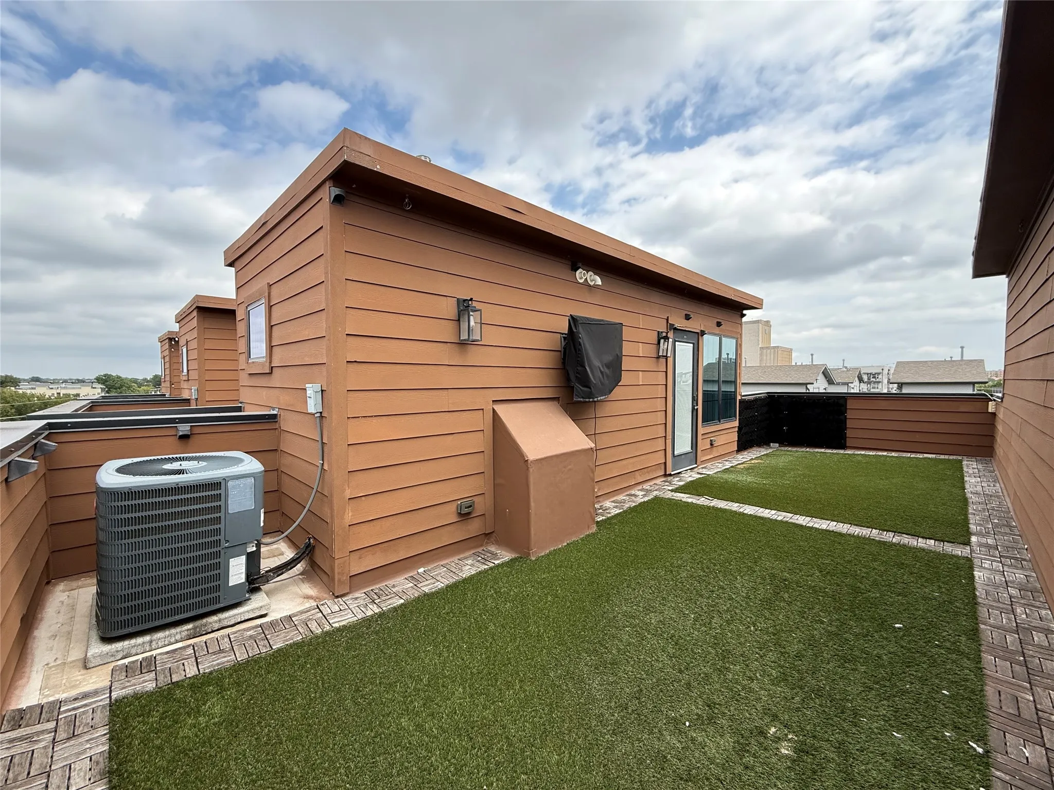 Rooftop deck featuring artificial turf, brick-patterned hardscaping, and access to an interior space via a glass-paneled door