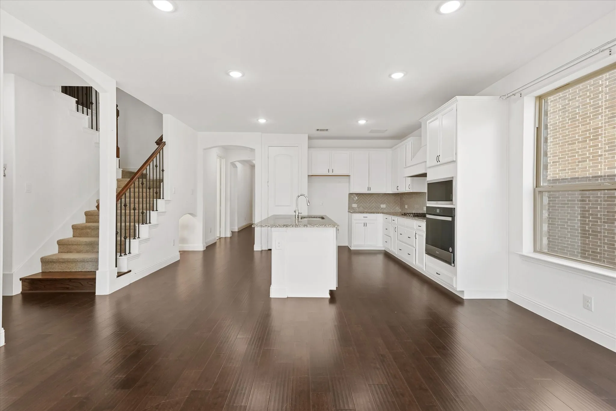 Expansive main living area with dark wood-finish flooring, recessed lighting, and a kitchen featuring white cabinetry, a built-in oven, and an island with a sink