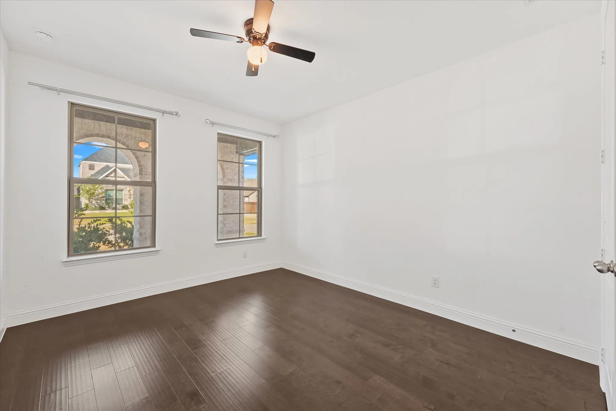 Wood-finish flooring, two large windows, white walls, and a ceiling fan with integrated lighting