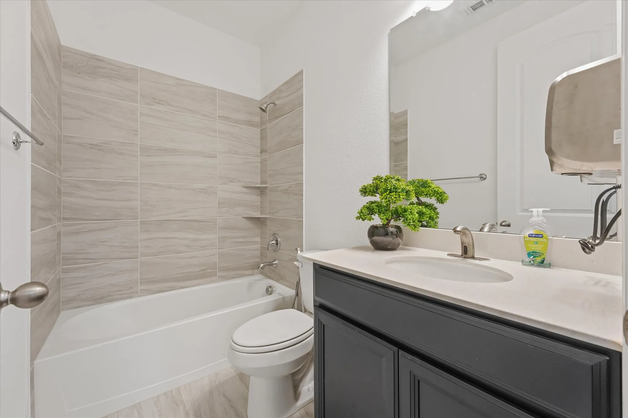 White bathtub with tile surround, built-in shelving, dark vanity with undermount sink, and wall-mounted mirror