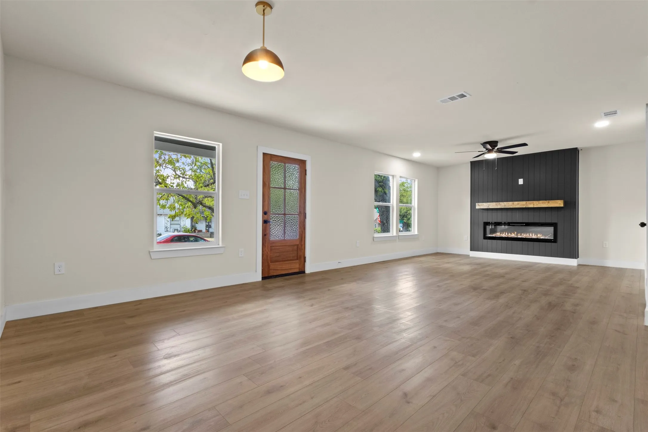 Expansive living area featuring light wood-finish flooring, a modern linear fireplace with a wood mantel, a black vertical slat accent wall, and recessed lighting