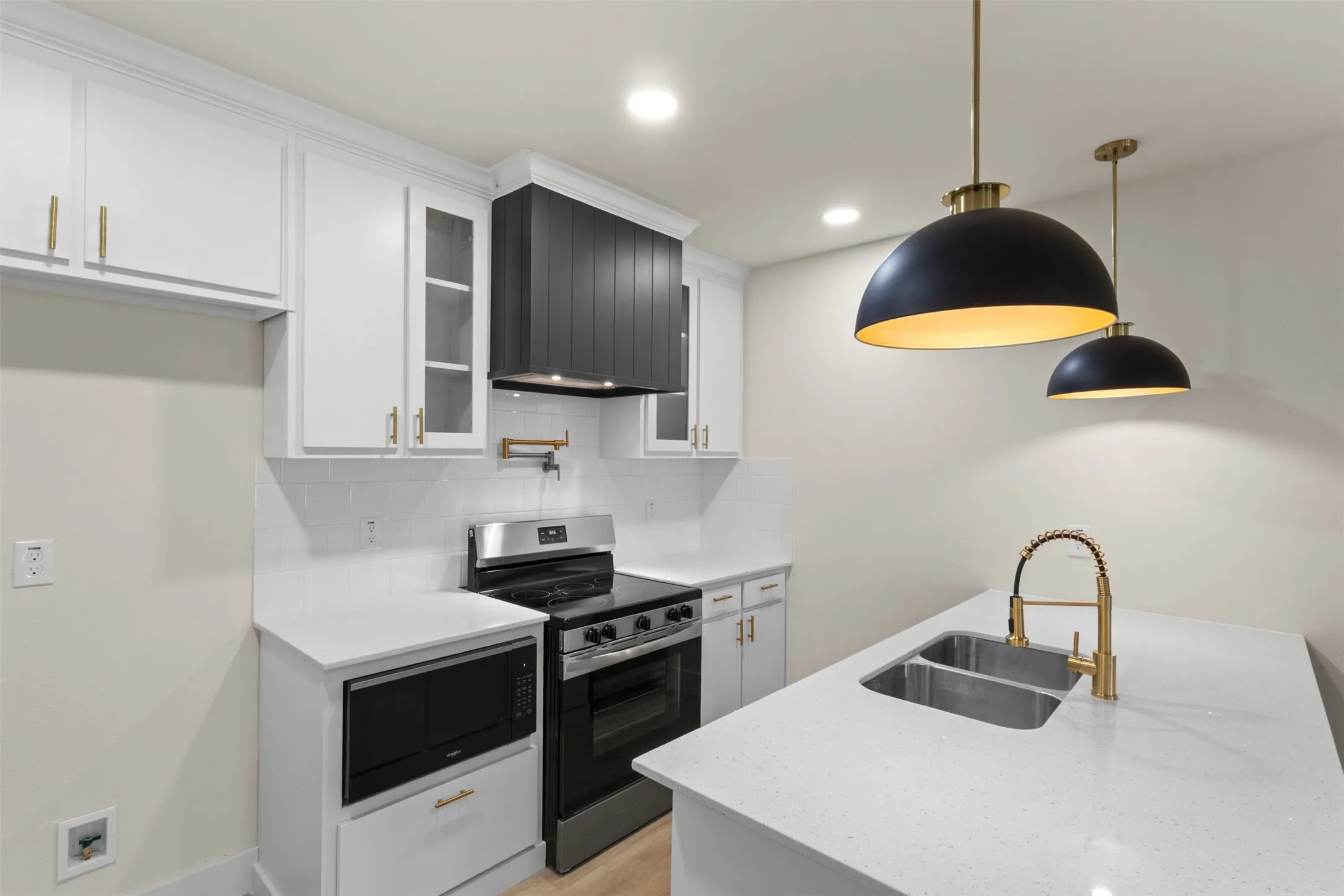 Kitchen featuring white cabinetry, a black range hood, stainless steel appliances, a double basin sink with a gold-finish faucet, and pendant lighting