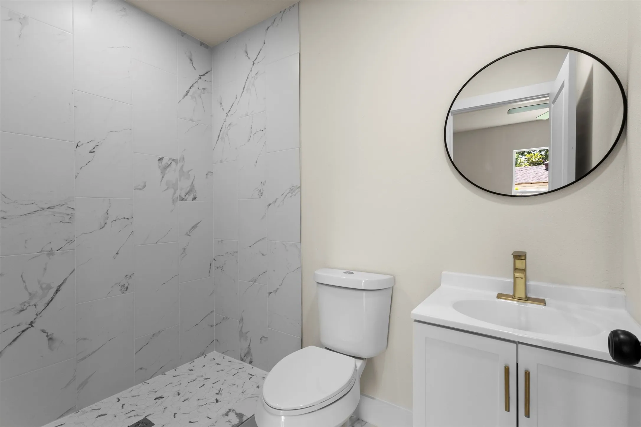 White vanity with gold-tone hardware, a circular mirror, and a walk-in shower with marble-style wall tiles and mosaic floor tiles