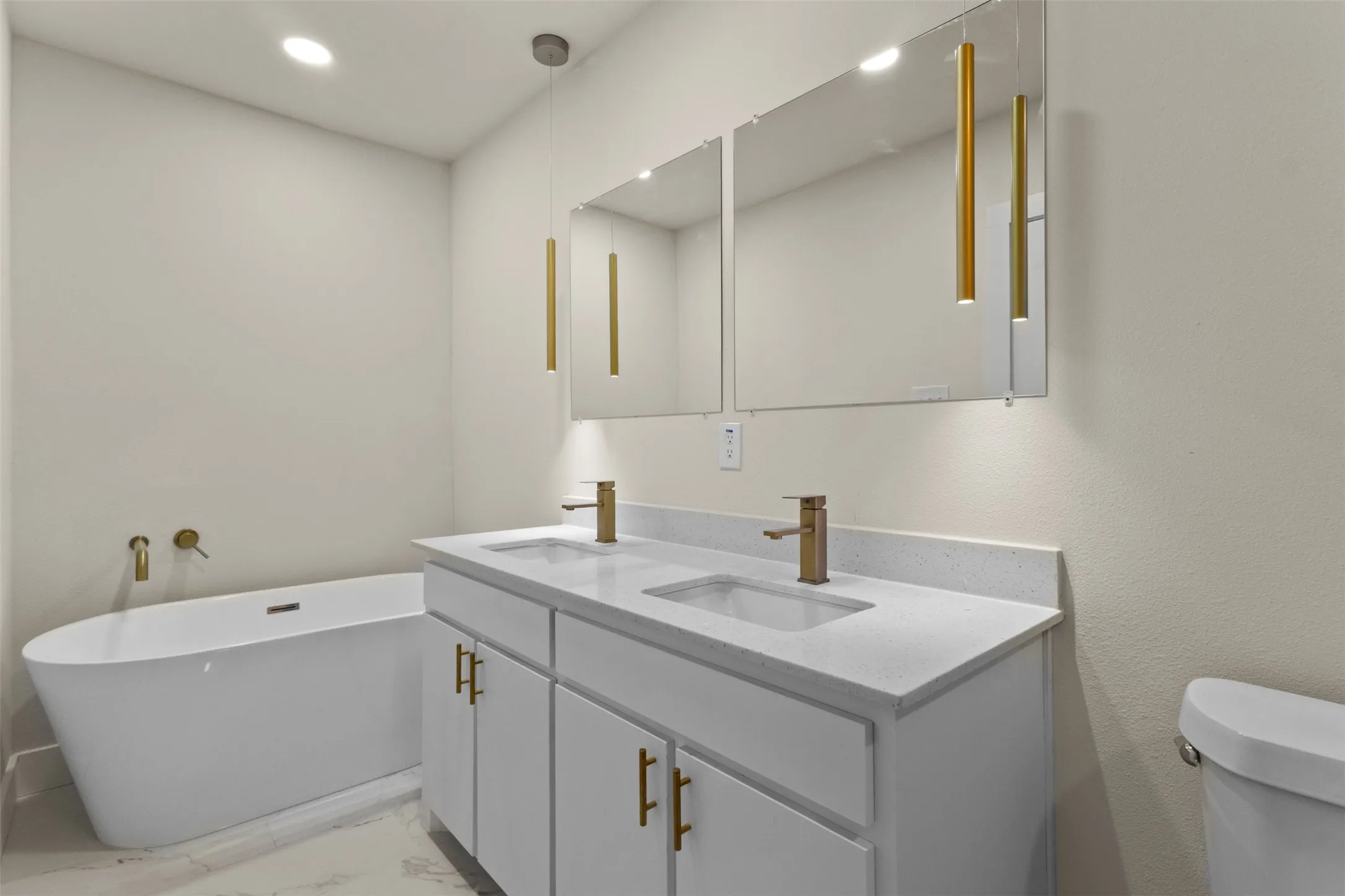 Freestanding soaking tub, dual vanity with undermount sinks, quartz countertops, brushed brass fixtures, and wall-mounted mirrors with integrated lighting