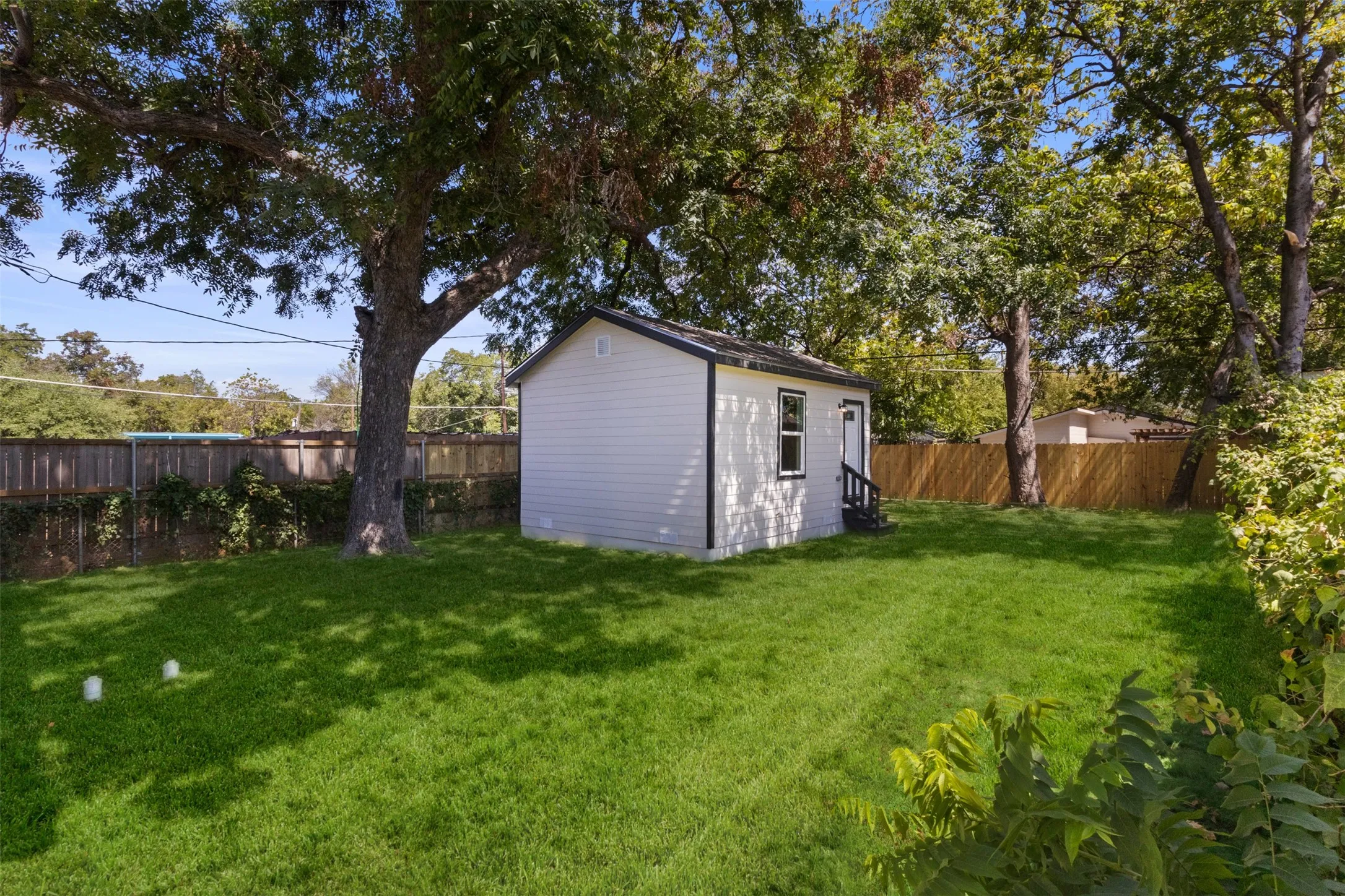 Expansive lawn featuring mature trees, a privacy fence, and an auxiliary structure with white siding and dark trim