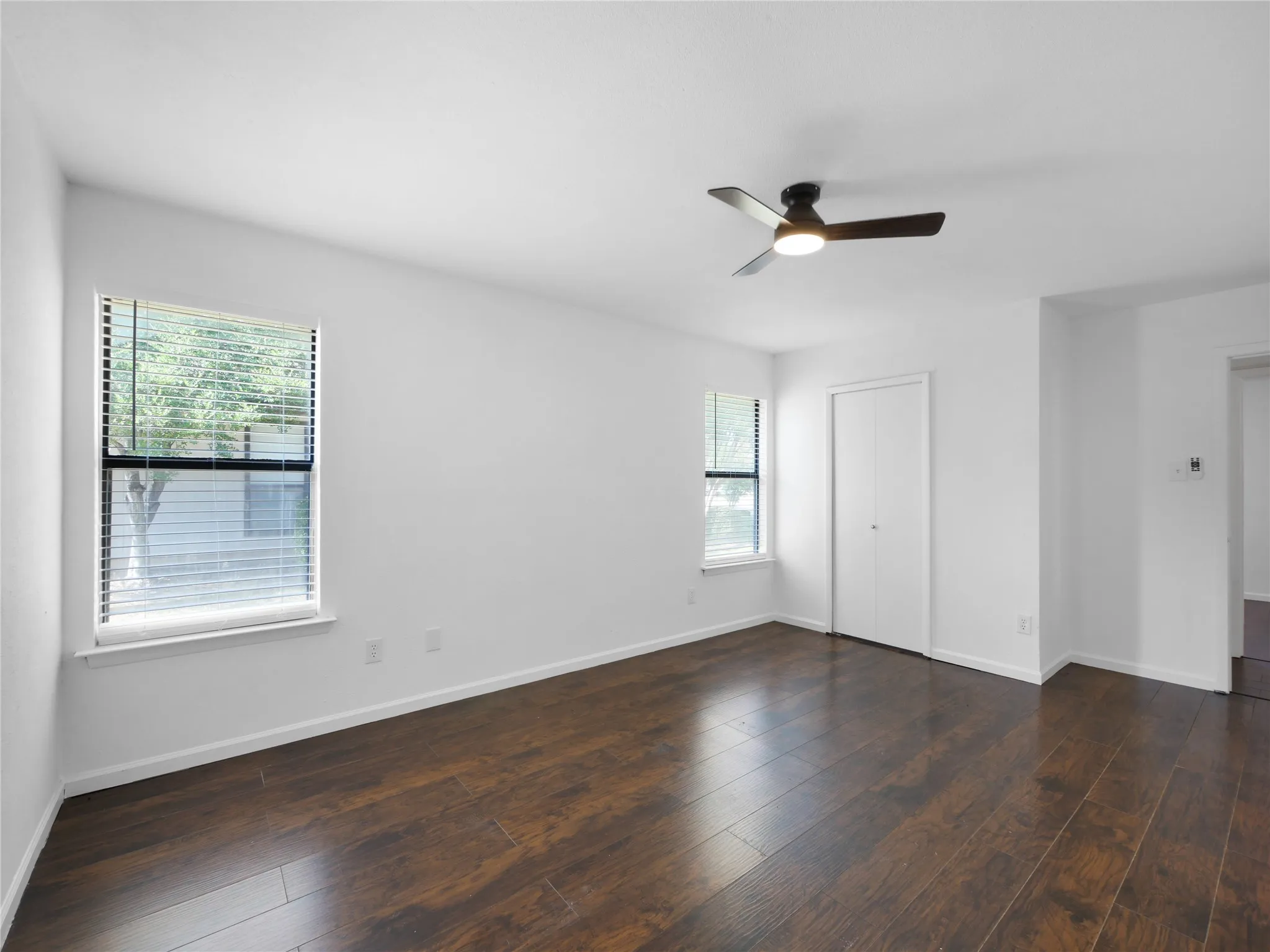 Expansive room featuring rich wood-finish flooring, a modern ceiling fan with integrated lighting, and two windows providing natural illumination