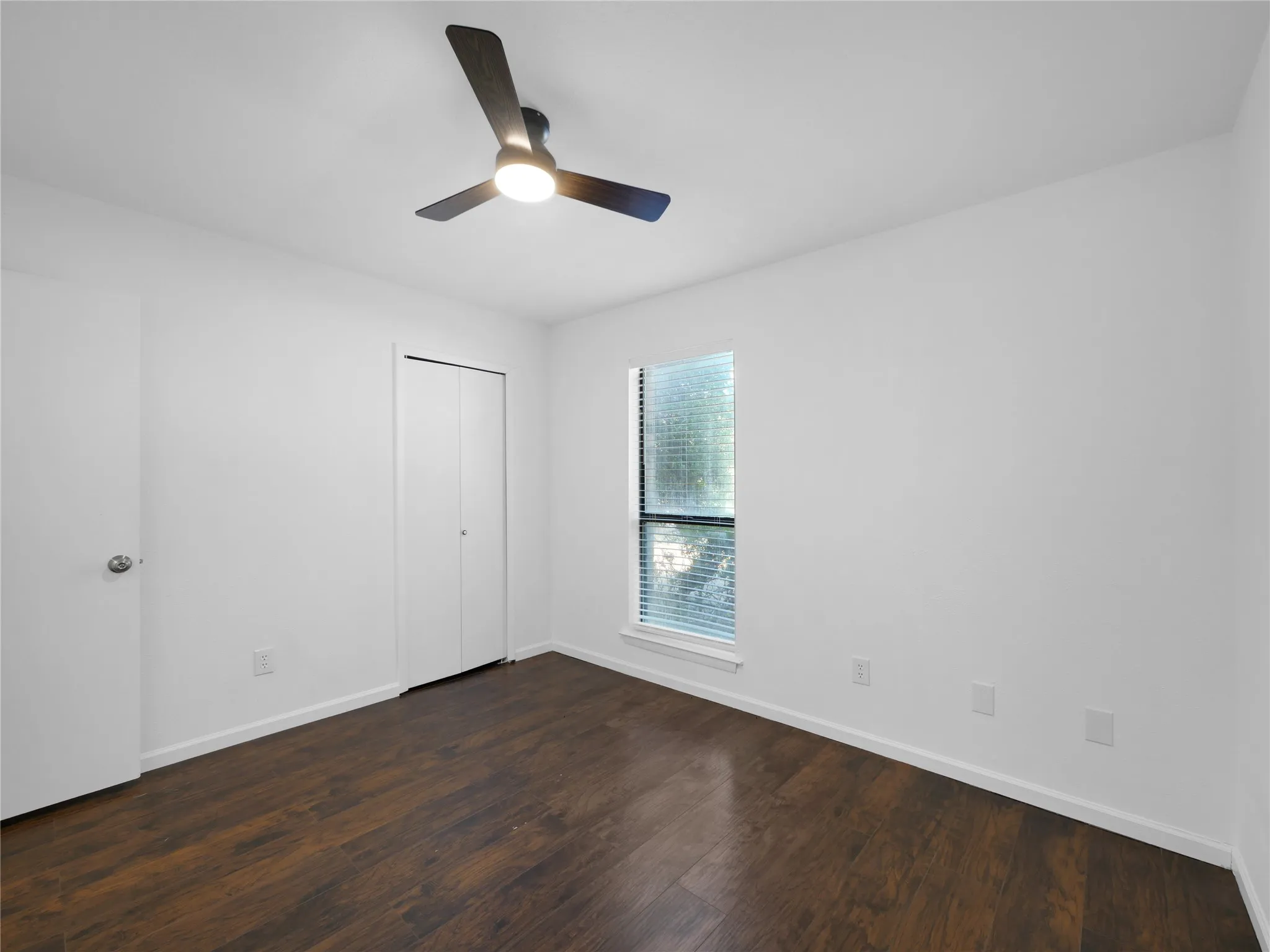 Dark wood-finish flooring, white walls, ceiling fan with integrated lighting, window with blinds, and white bi-fold closet doors