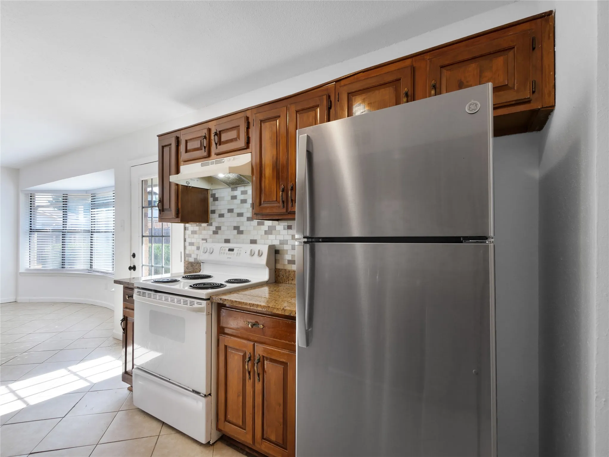 Kitchen featuring wood cabinetry, a mosaic tile backsplash, a white electric range, granite-finish countertops, and a stainless steel refrigerator