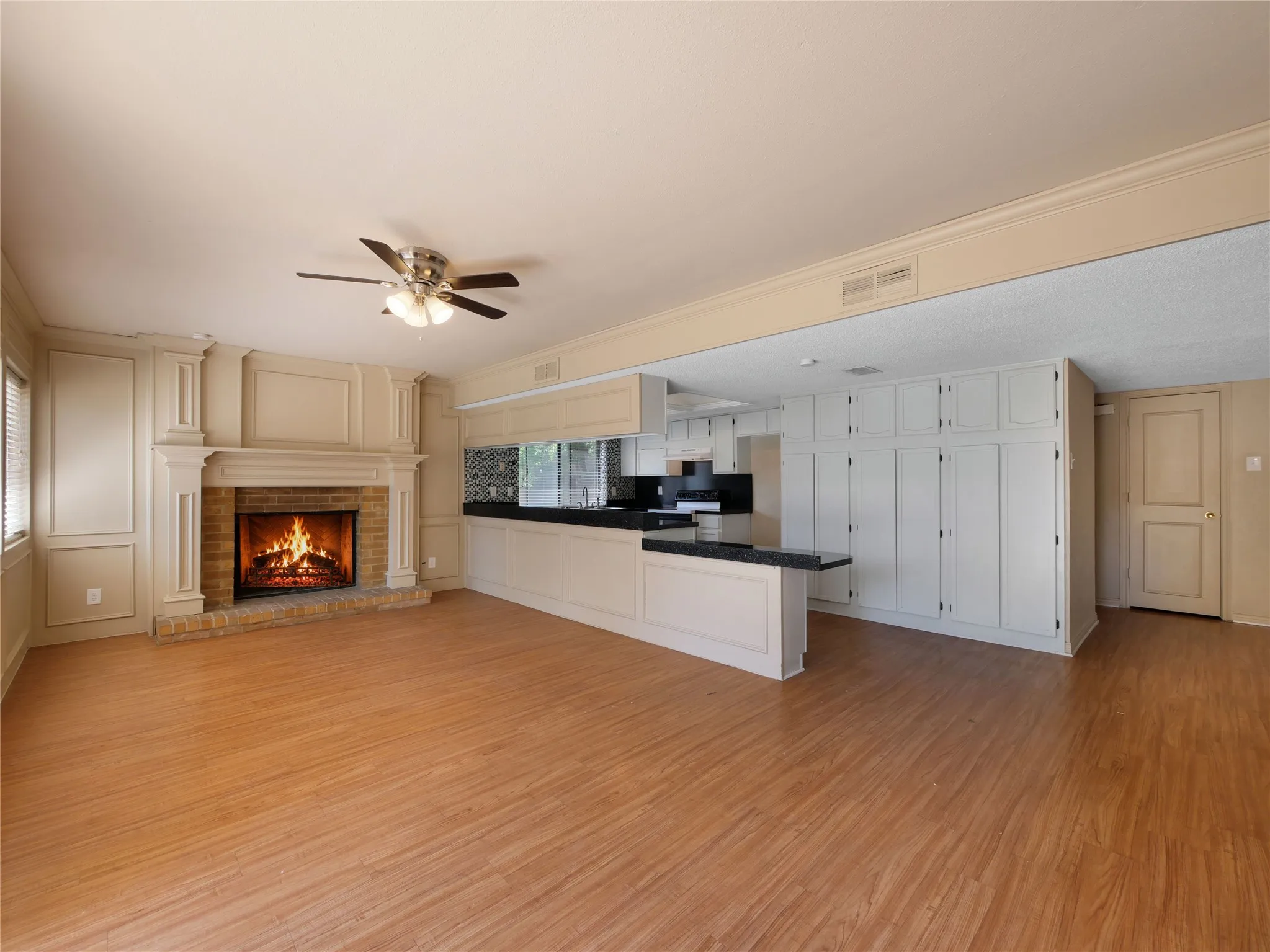 Spacious room featuring wood-finish flooring, a brick fireplace with a decorative mantel, a ceiling fan, and built-in white cabinetry with dark countertops