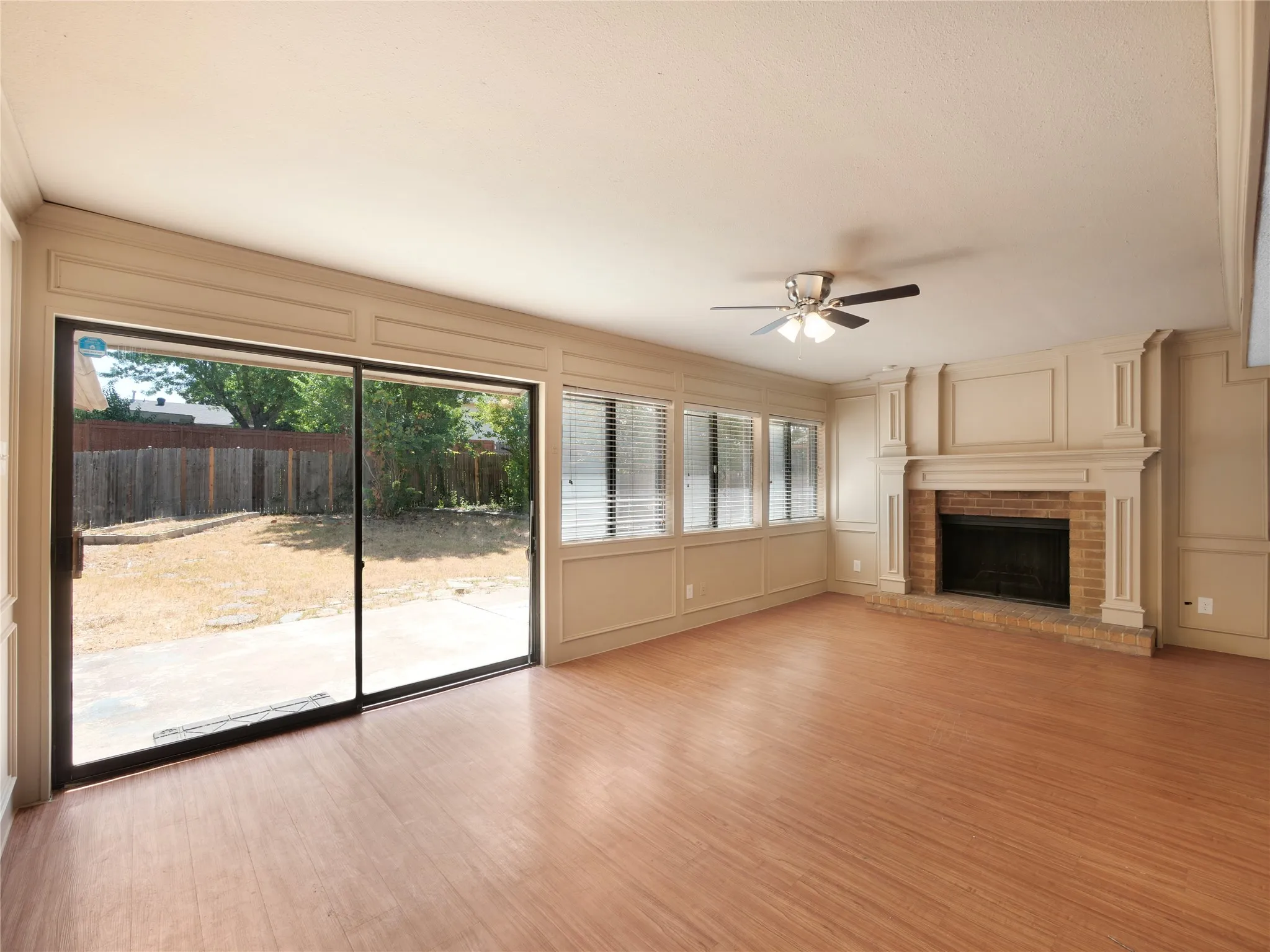 Spacious room featuring wood-finish flooring, a brick fireplace with an ornate mantel, multiple windows with blinds, a ceiling fan with light, and a sliding glass door to the backyard