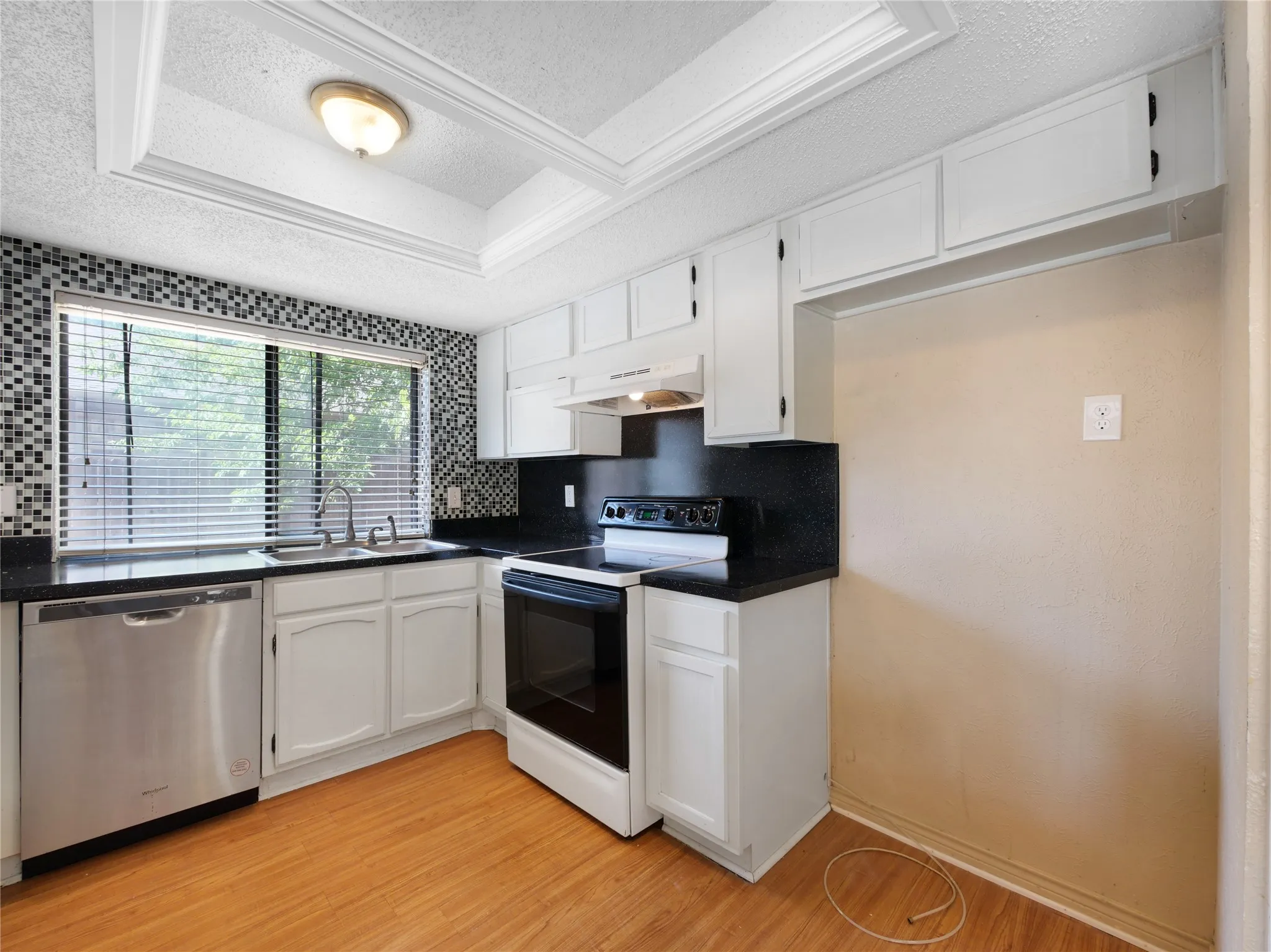 Kitchen featuring white cabinetry, black countertops, stainless steel dishwasher, mosaic tile backsplash, and wood-finish flooring