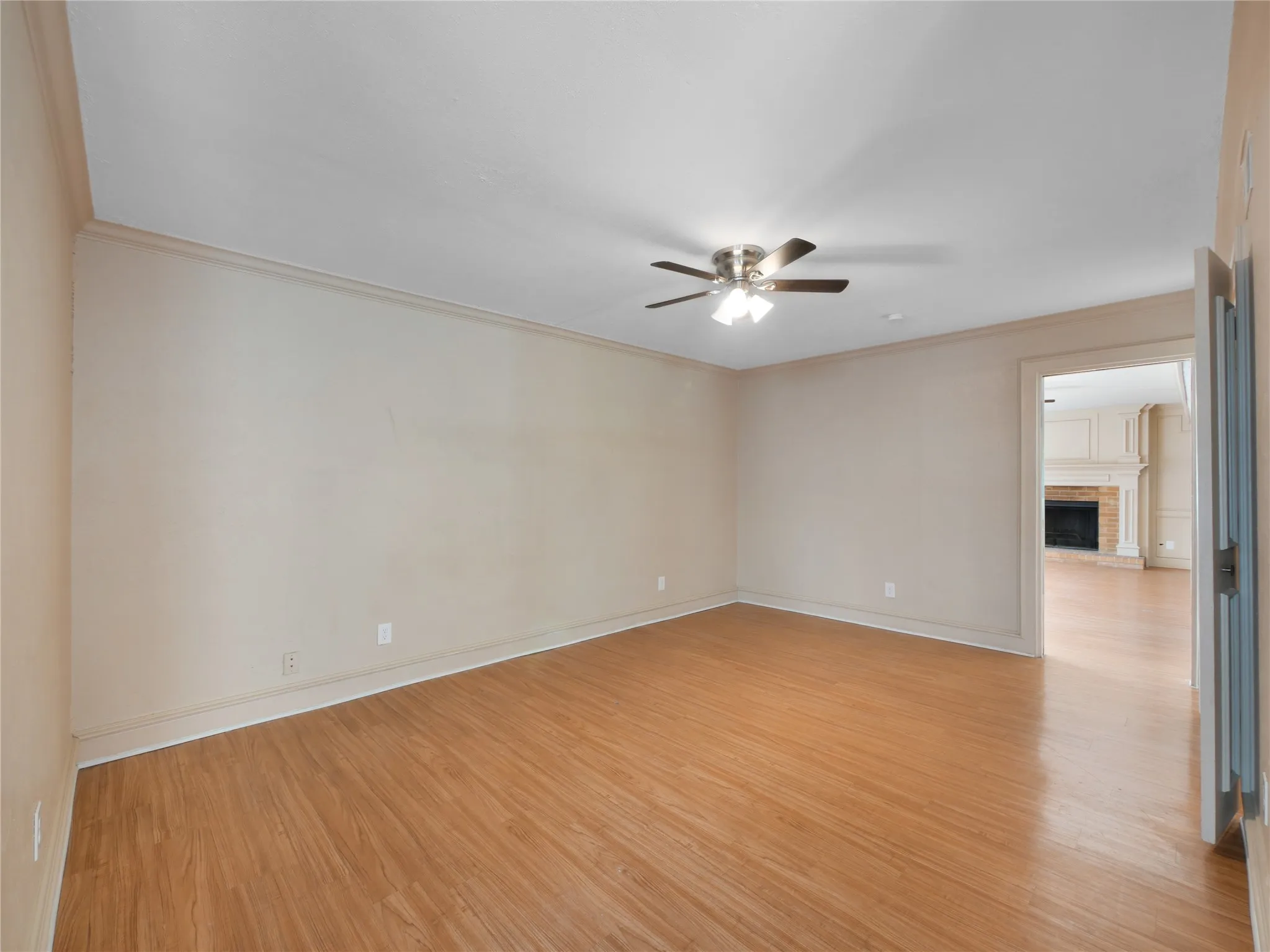 Wood-finish flooring, ceiling fan with integrated lighting, crown molding, and an open doorway leading to a room with a fireplace