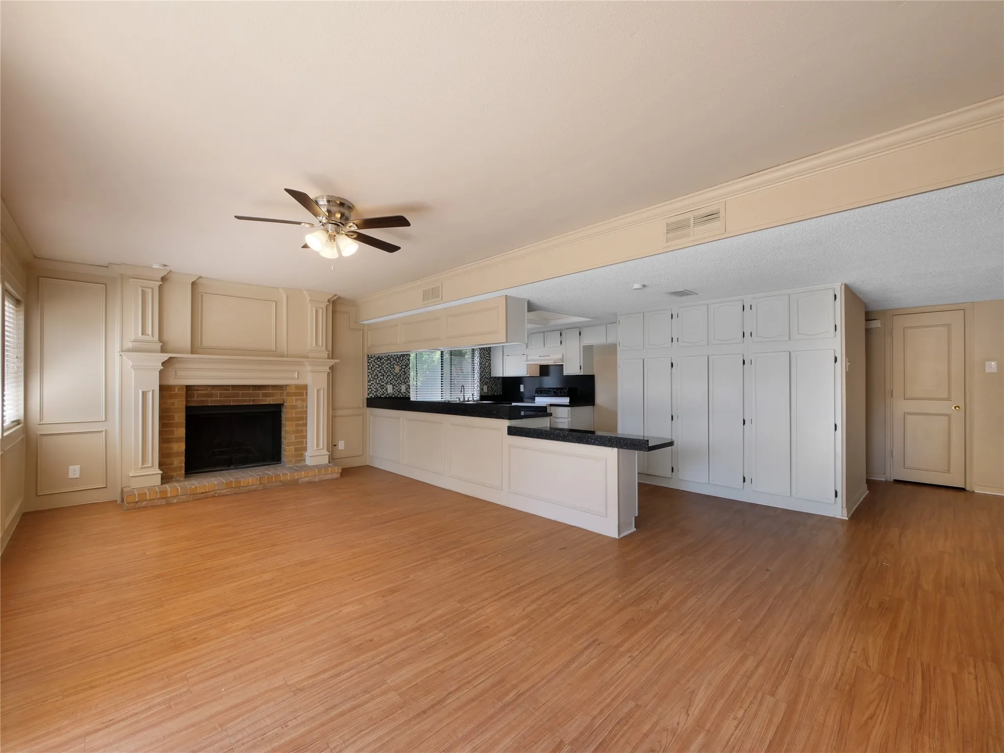Wood-finish flooring throughout, brick-surround fireplace with detailed mantel, ceiling fan with integrated lighting, kitchen featuring extensive white cabinetry, and a dark stone countertop with breakfast bar seating