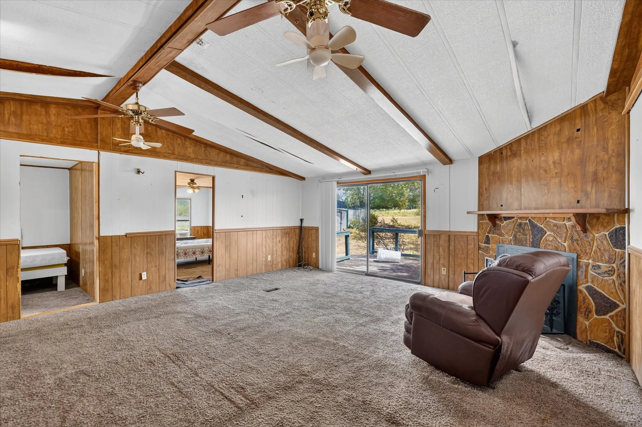 Vaulted ceiling with exposed wood beams, stone-faced fireplace, wood paneling, sliding glass door, and ceiling fans