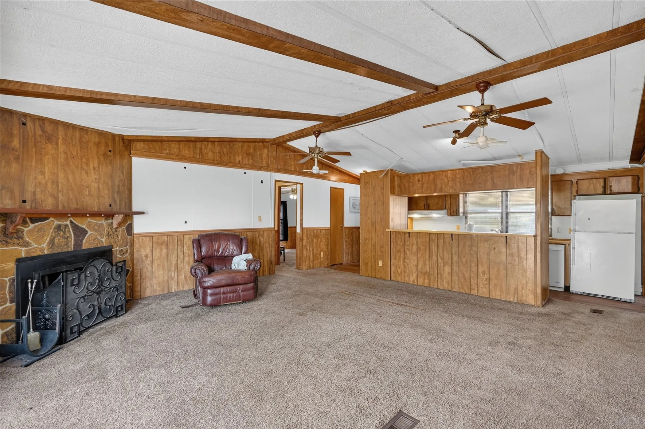 Exposed ceiling beams, two ceiling fans, stone-finish fireplace, wood-finish wall panels, and a kitchen pass-through window
