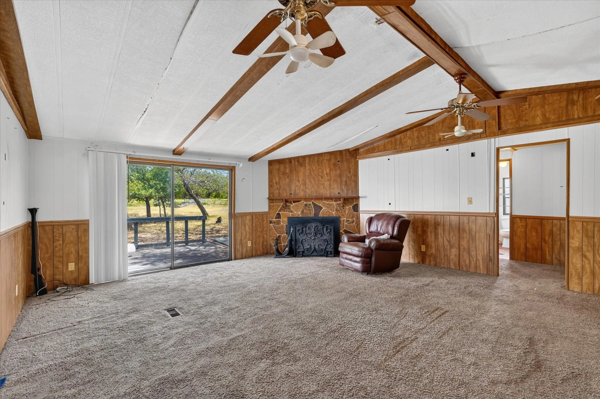 Vaulted ceiling with exposed wood beams, stone-faced fireplace, ceiling fans, and sliding glass door