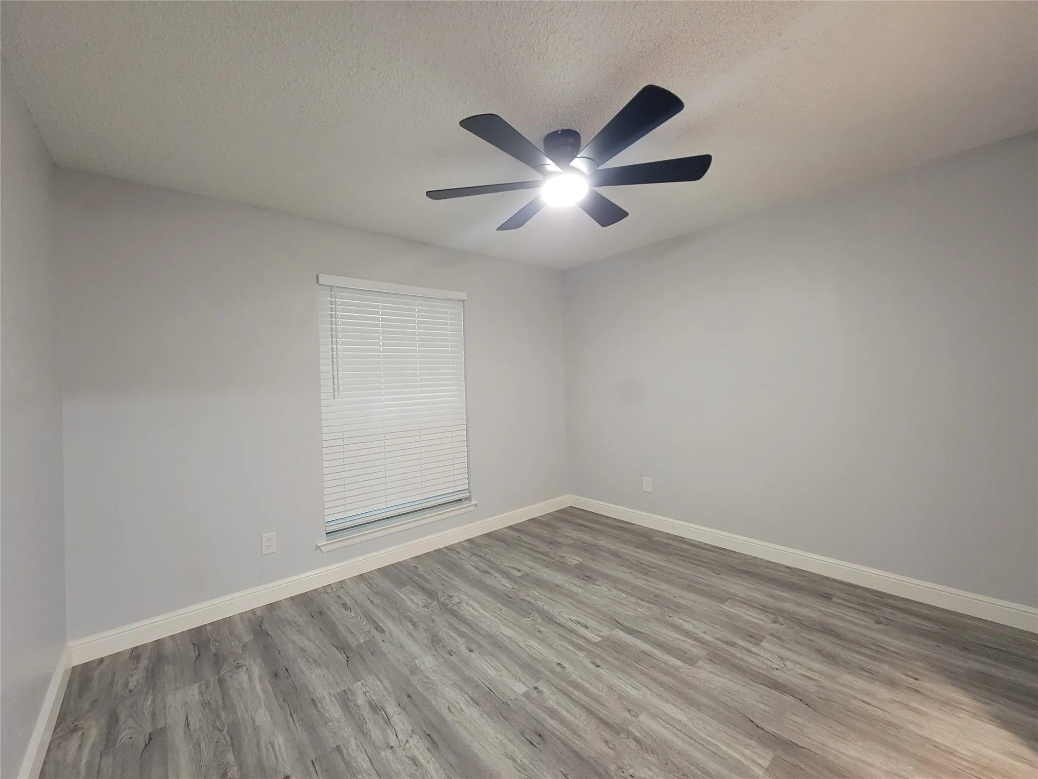 Spacious room featuring wood-finish flooring, a contemporary ceiling fan with integrated lighting, and a window with blinds