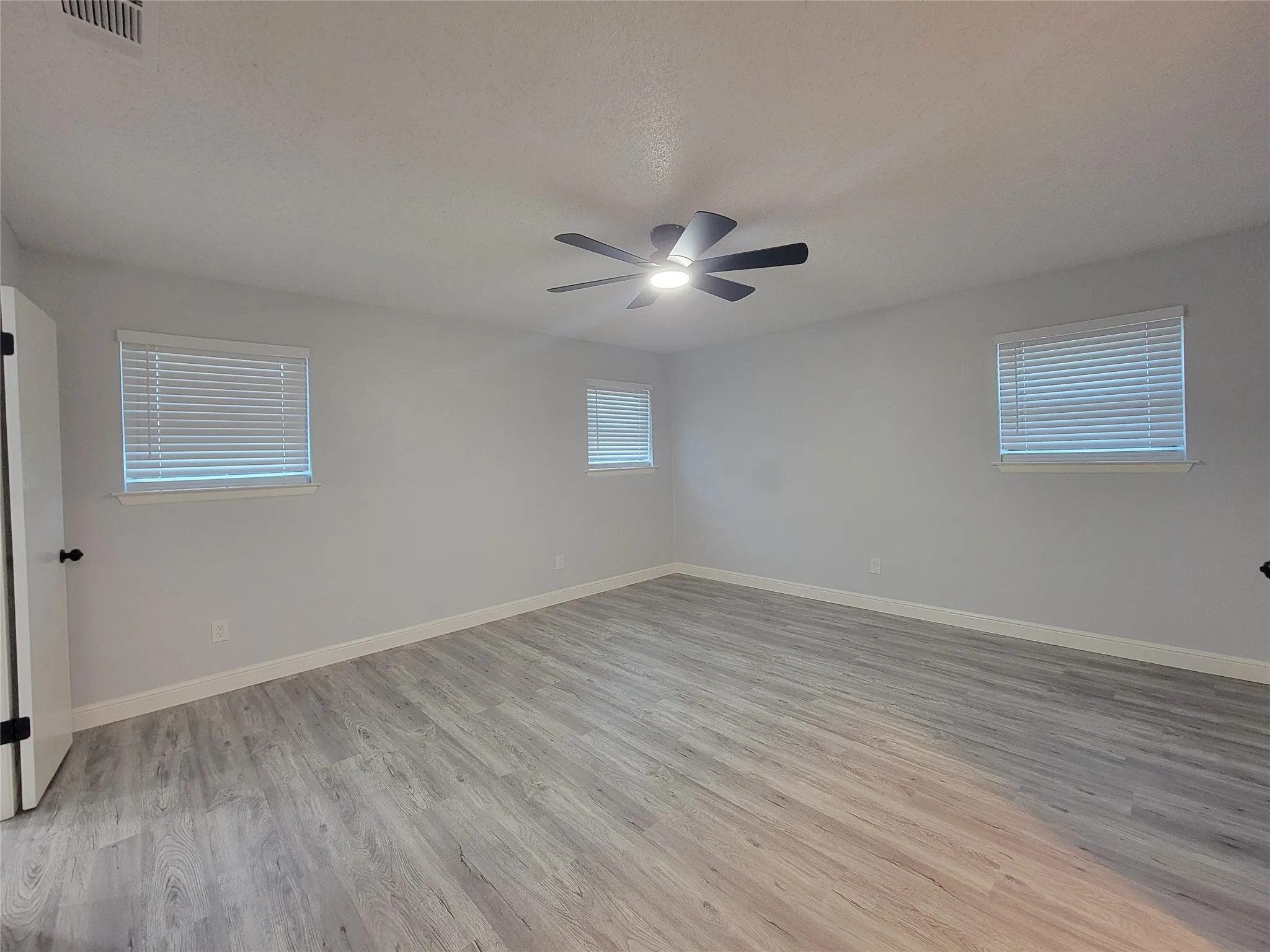 Spacious room featuring light-colored walls, wood-finish flooring, a ceiling fan with integrated lighting, and multiple windows with blinds