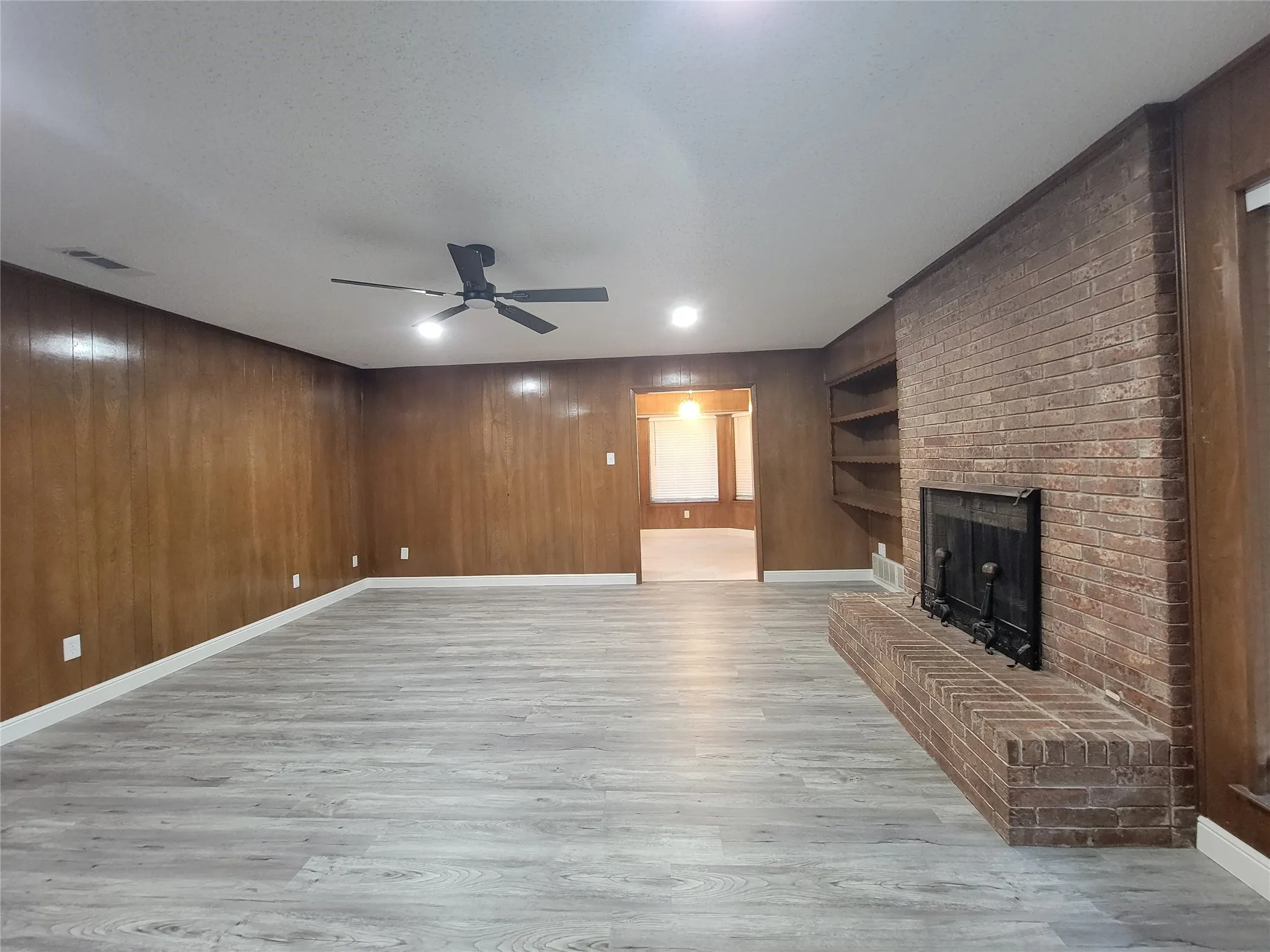 Spacious room featuring wood-finish wall panels, light wood-finish flooring, a brick fireplace with hearth, built-in shelving, and a ceiling fan with integrated lighting
