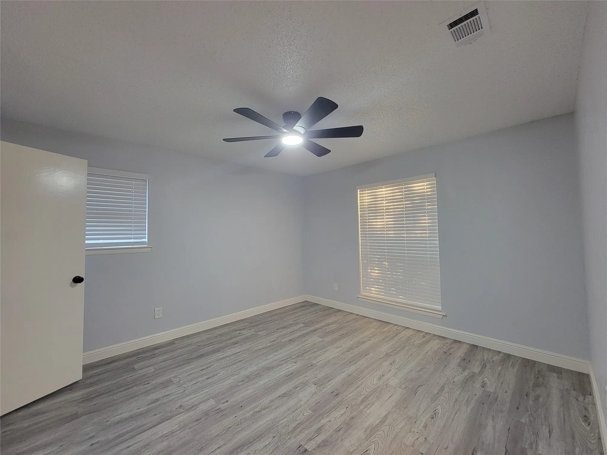 Light gray walls, wood-finish flooring, ceiling fan with integrated lighting, and two windows with blinds