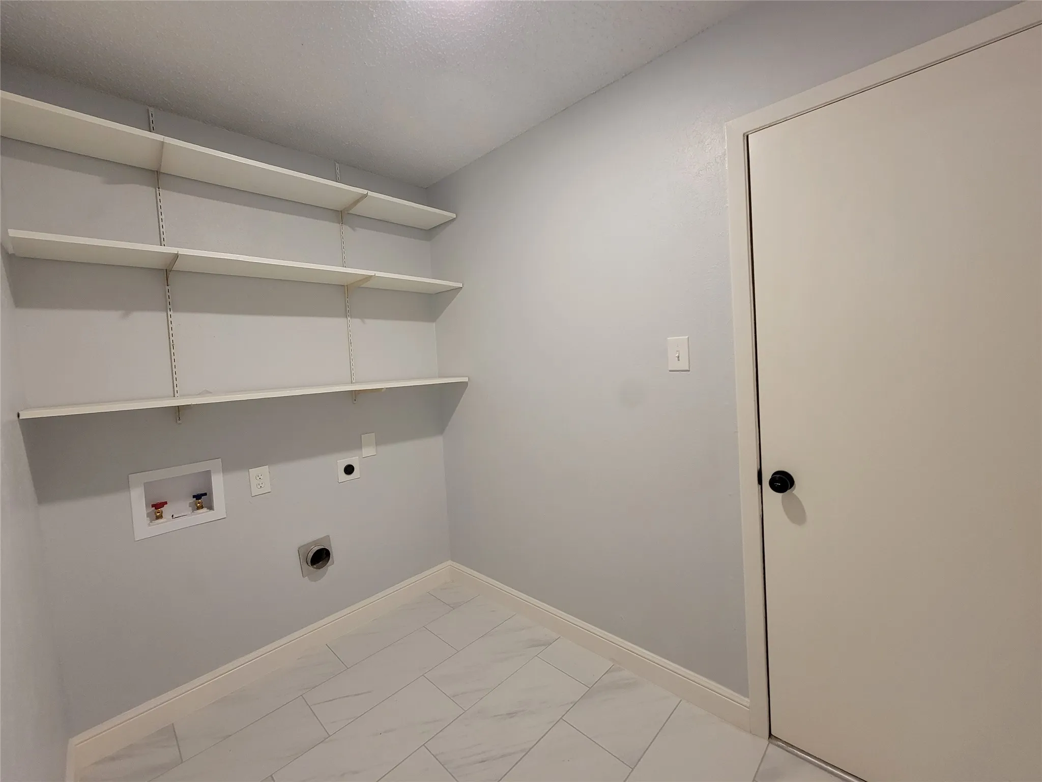 Laundry area featuring light-toned tile flooring, three adjustable wall-mounted shelves, and utility hookups