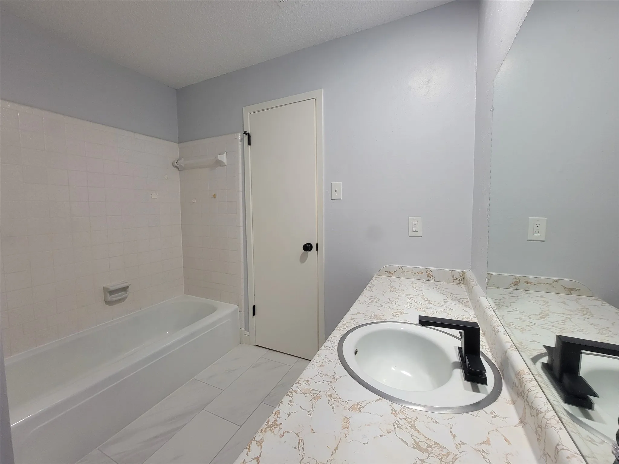 Bathroom featuring a white bathtub, white tiled shower surround, patterned countertop, and a vanity with an integrated sink and black faucet