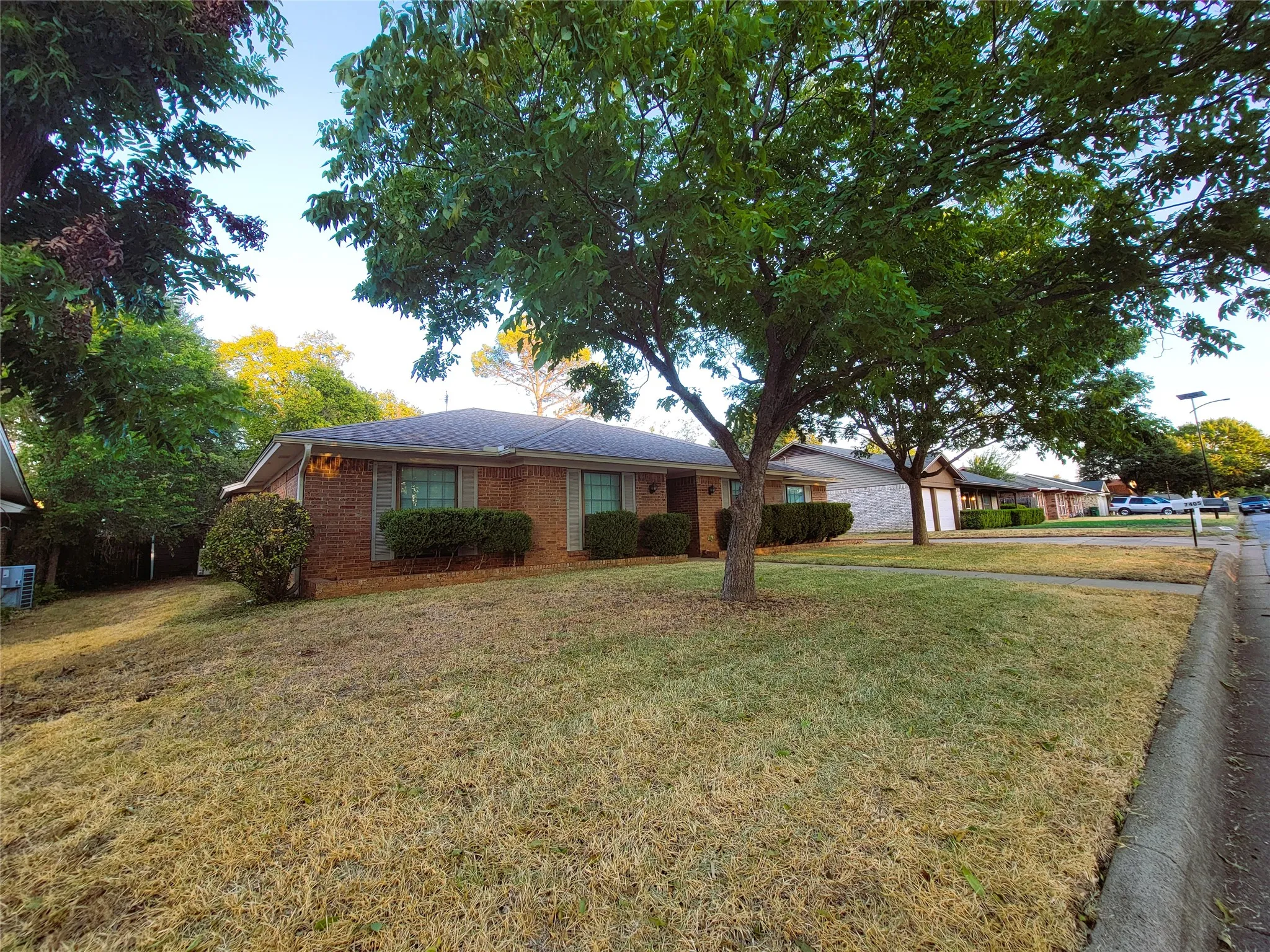 Brick exterior, pitched roof, mature trees, manicured shrubs, and a sidewalk