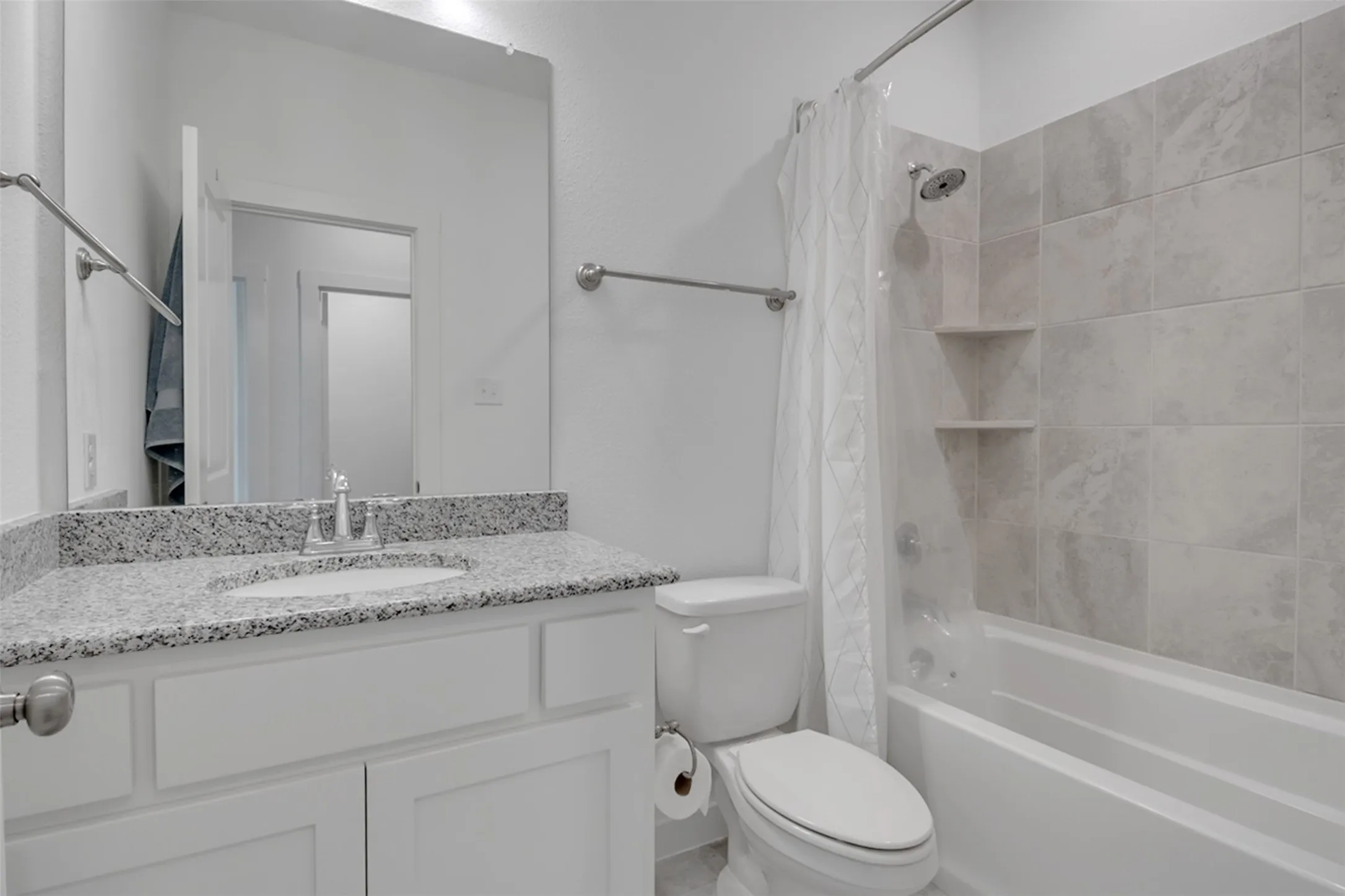 Vanity with a granite countertop and undermount sink, white cabinetry with silver hardware, and a tub-shower combination with light gray wall tile and built-in shelving