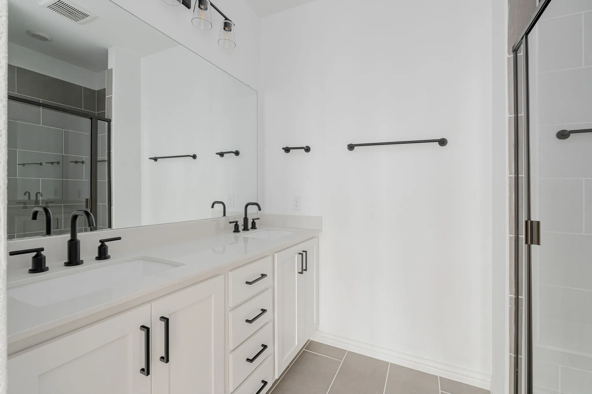 Fresh and contemporary primary bath features an extended dual-vanity with sleek countertops, modern matte-black fixtures, oversized mirror, and a spacious walk-in shower with a glass enclosure.