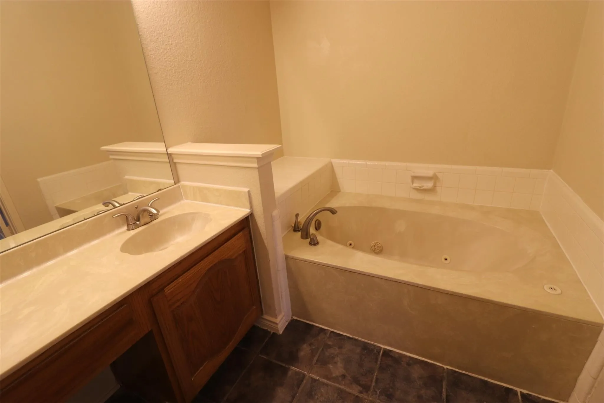Built-in jetted tub with tile surround, single vanity with integrated sink, dark wood-finish cabinetry, and large wall-mounted mirror