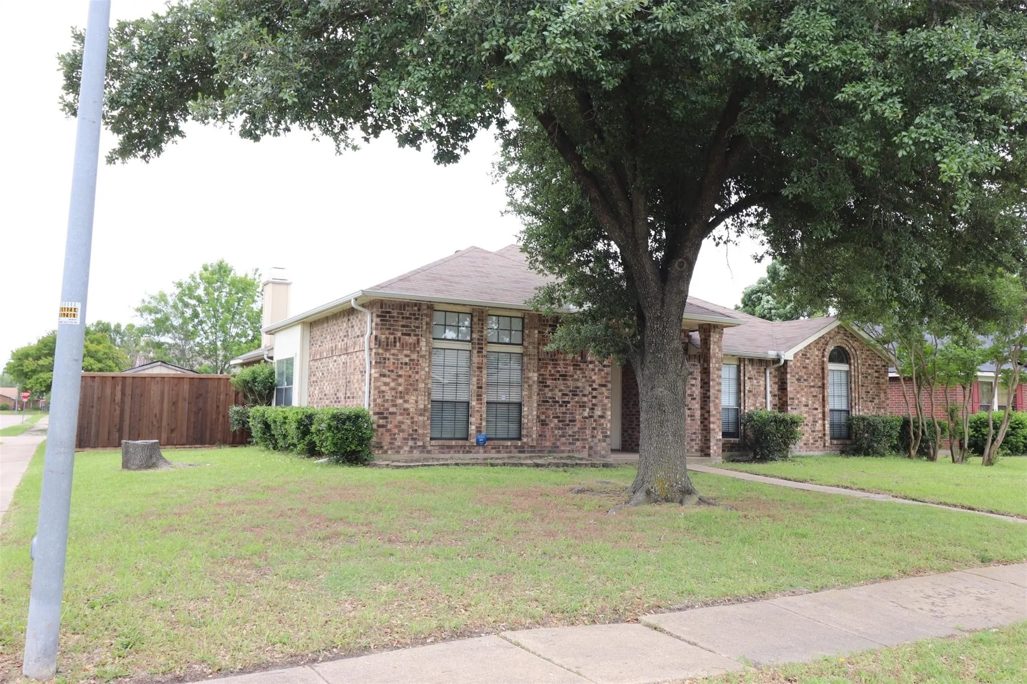 Brick exterior, mature front yard tree, landscaped shrubbery, fenced backyard, and a concrete sidewalk