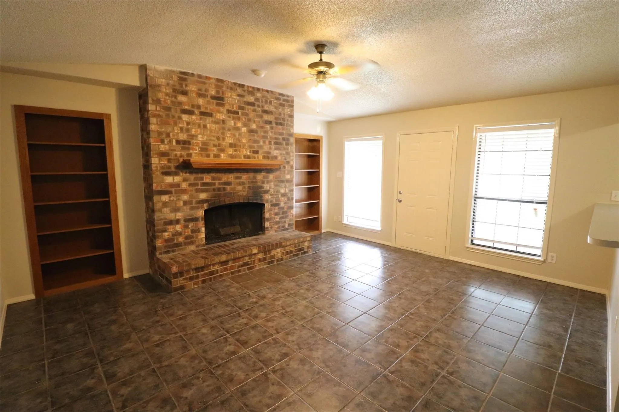 Brick fireplace with a wood mantle, built-in shelving, tile flooring, and a ceiling fan