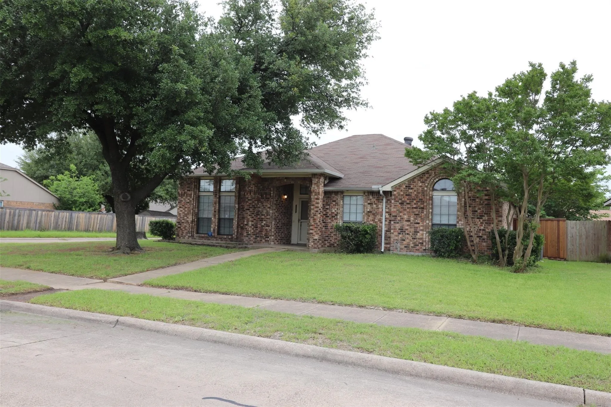 Brick exterior with a brown shingle roof, mature trees, and a concrete sidewalk
