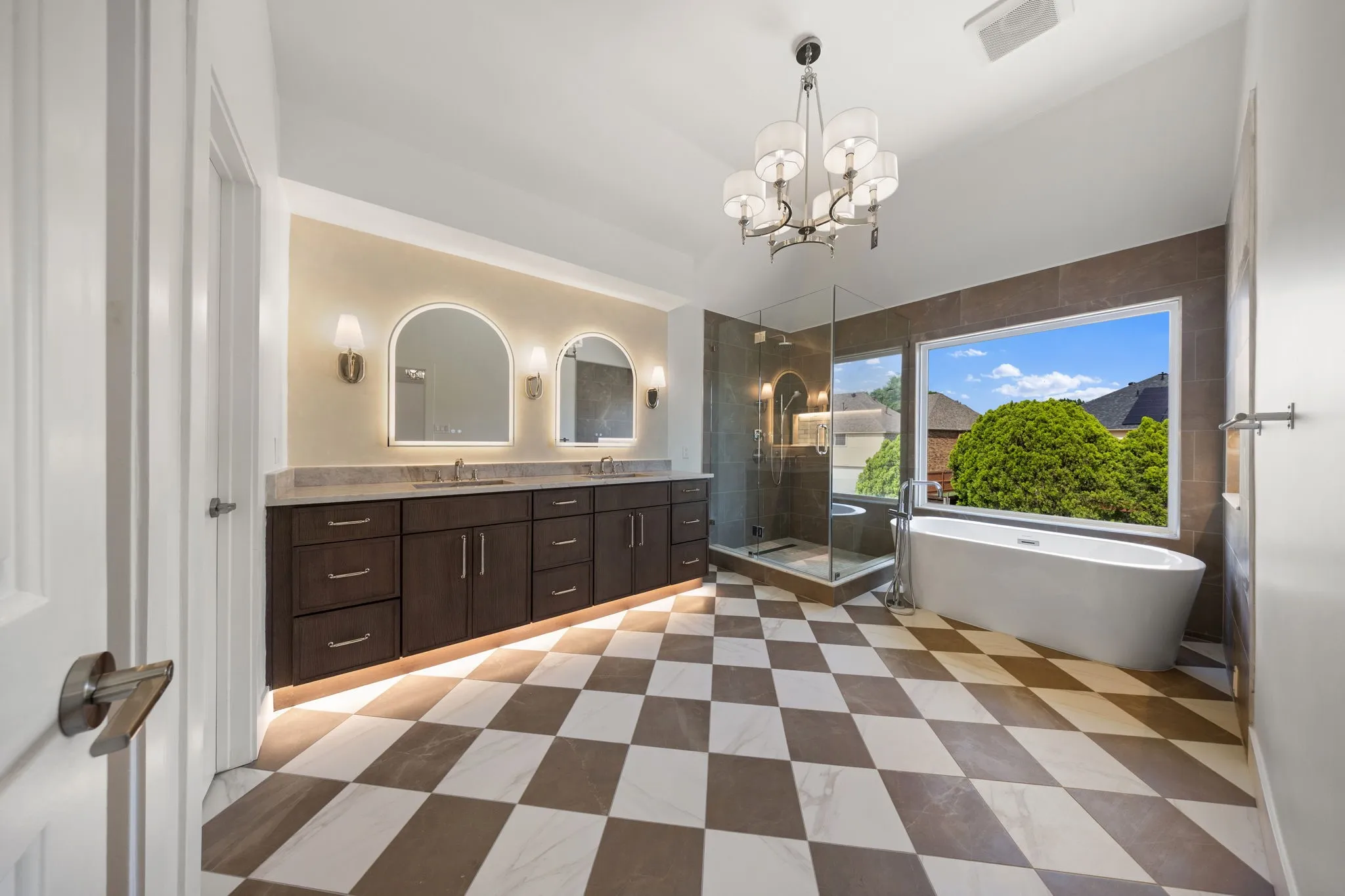 Spacious bathroom featuring checkered tile flooring, a freestanding soaking tub, a frameless glass shower enclosure, a dual vanity with undermount sinks, and illuminated arched mirrors