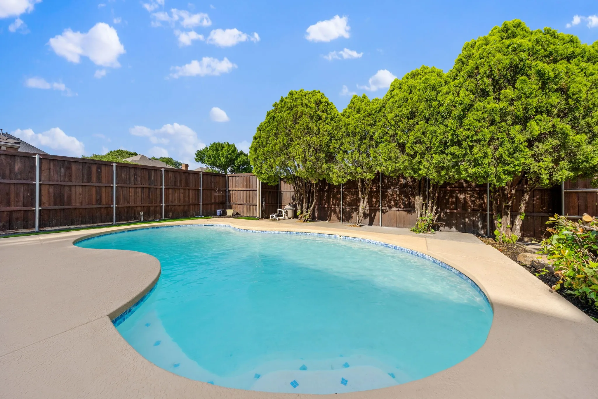 Curved swimming pool featuring a light-colored concrete deck, blue waterline tiling, and a surrounding privacy fence