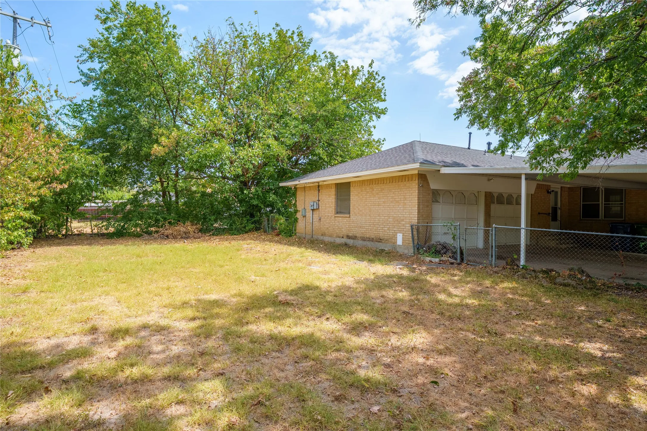 Expansive lawn area with mature perimeter trees, a brick exterior, and a covered carport with an attached chain-link fence