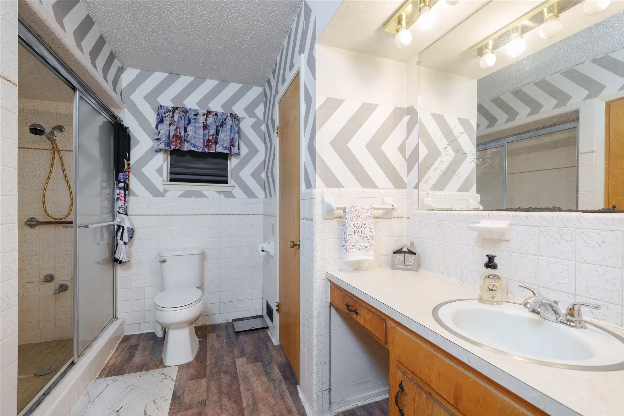 Shower with glass enclosure, white tile walls, wood-finish flooring, and a vanity with an integrated sink and mirror
