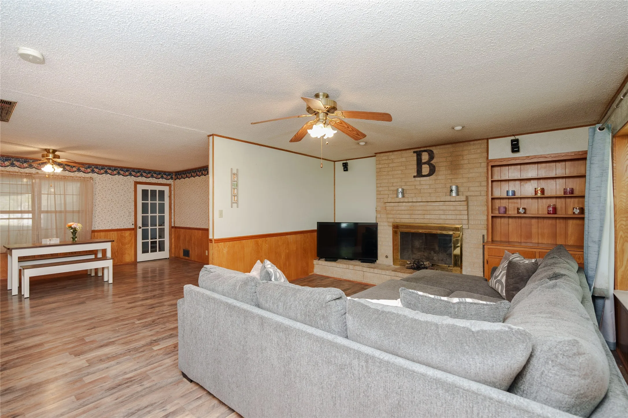 Brick fireplace with a brass-toned insert, wood-finish flooring, built-in shelving, paneled wainscoting, and a ceiling fan