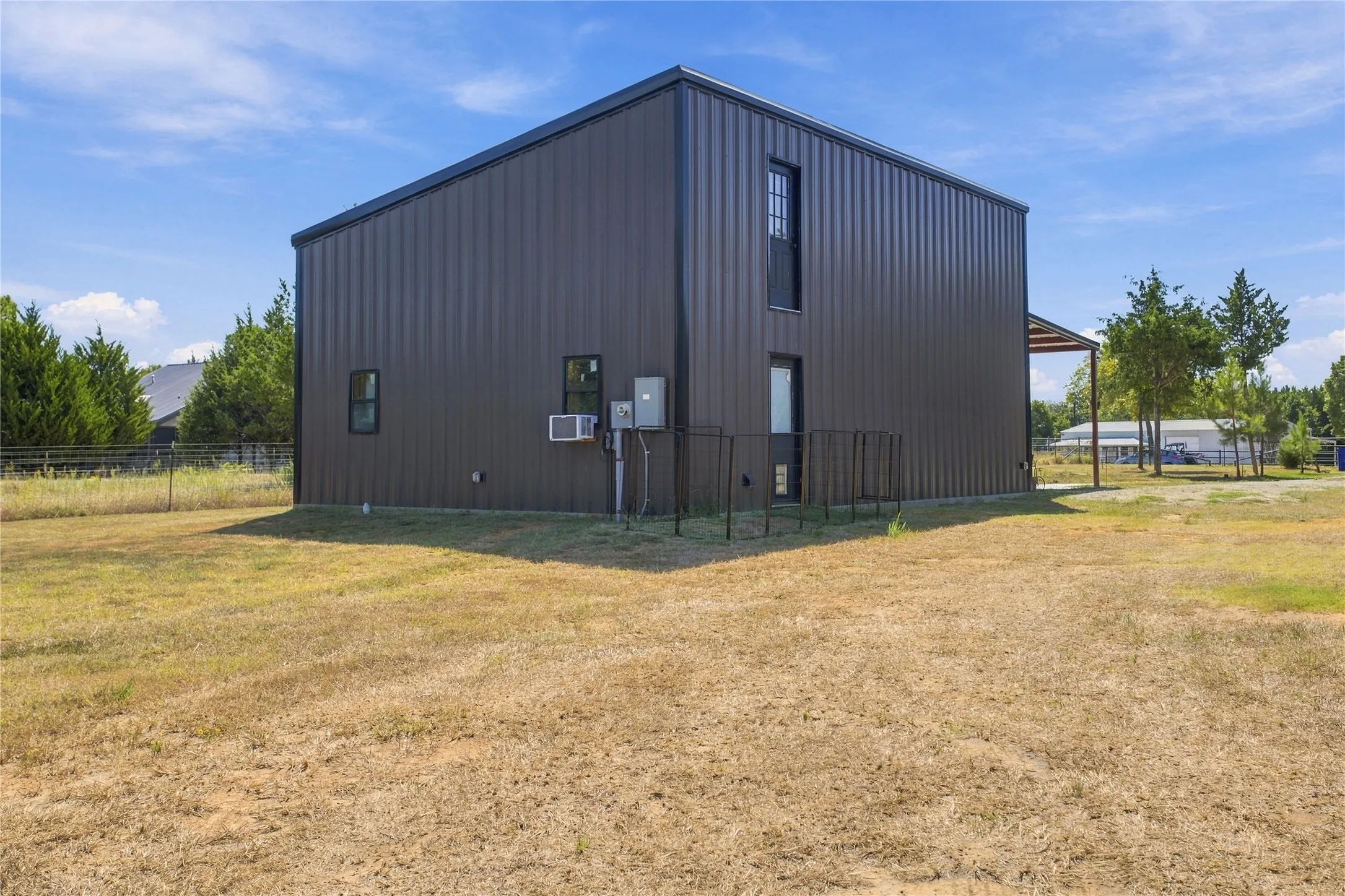 Metal-sided structure with a covered entry, multiple windows, and a black security fence