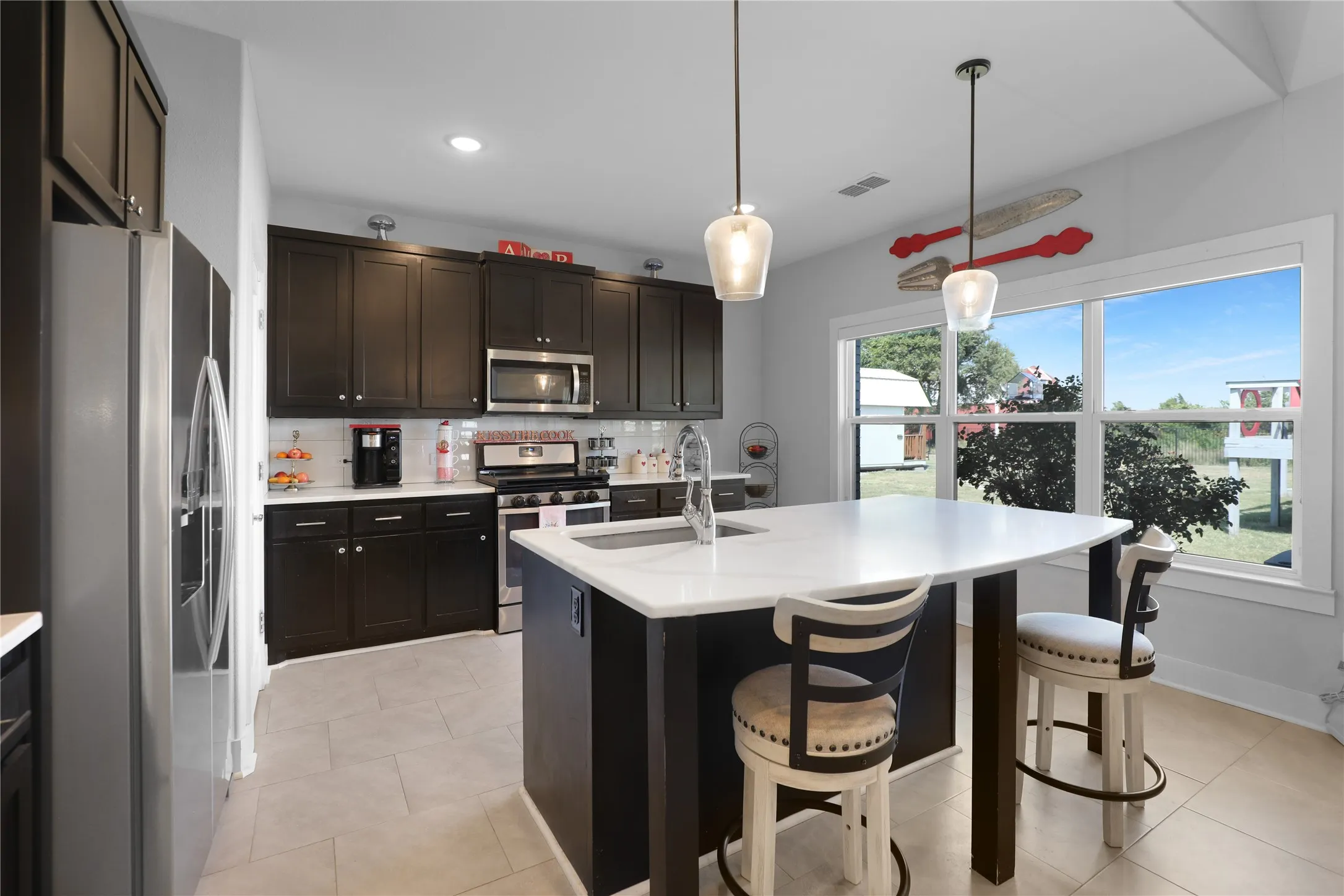 Kitchen featuring dark wood cabinetry, a white subway tile backsplash, stainless steel appliances, a center island with a white countertop and integrated sink, and light-toned tile flooring