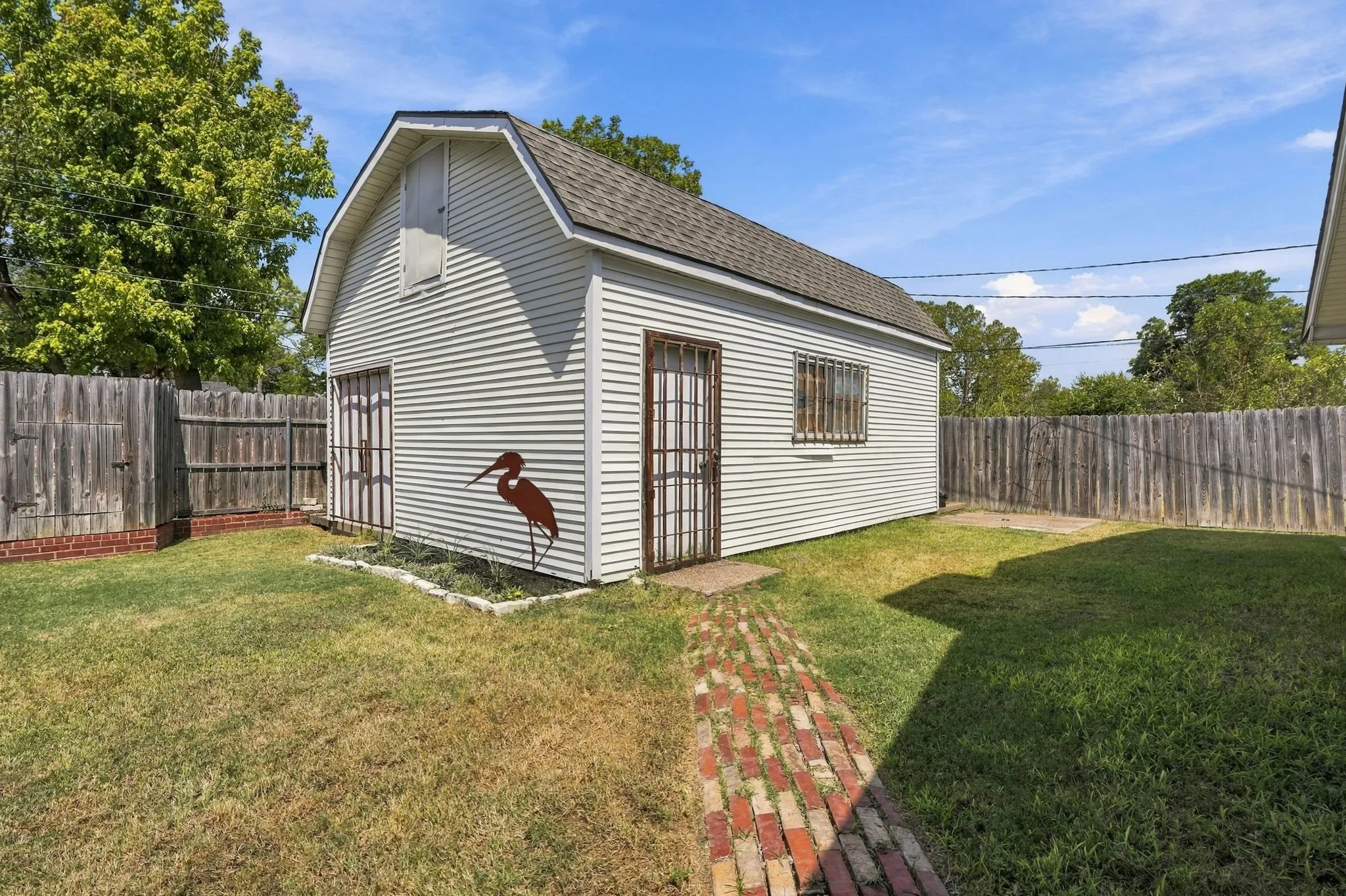 Detached utility building with gambrel roof, two entry doors, security bars on windows, brick pathway, and a perimeter fence