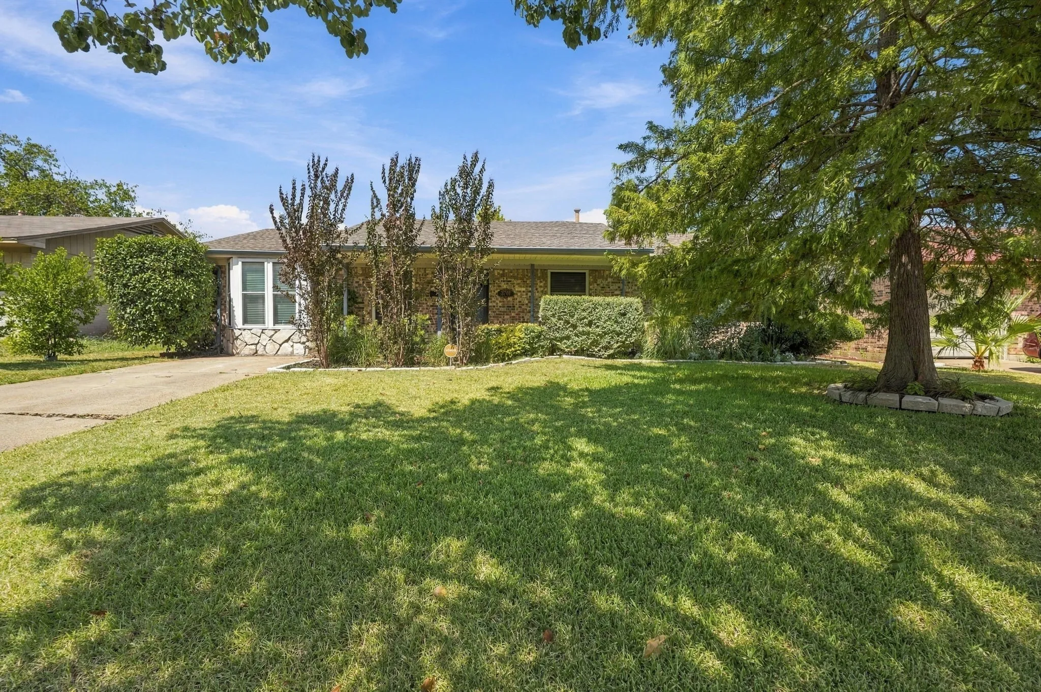 Stone veneer facade with a covered entry, mature landscaping, expansive front lawn, and a concrete driveway